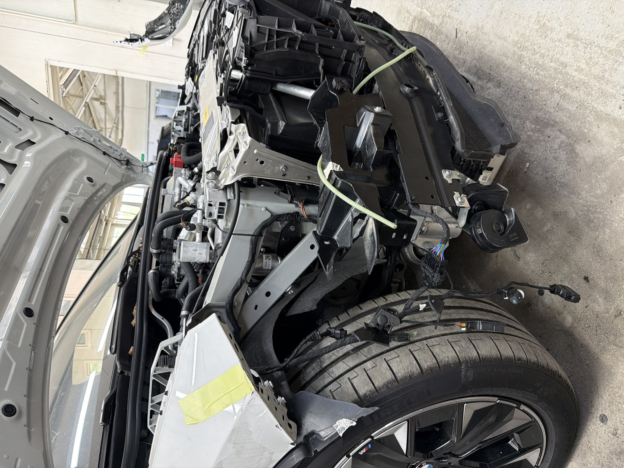 The image captures a partially disassembled white car in what appears to be an automotive repair shop or garage. The car's front end is extensively opened, with the bumper and possibly other body panels removed, exposing the engine bay components, structural elements, wiring harnesses, and hoses. This suggests the car is undergoing significant repair, maintenance, or reconstruction.
In the foreground, a prominent car wheel with a tire is visible, featuring a multi-spoke design and a BMW logo in the center, along with an 'M' logo on the rim, indicating it's likely a BMW M-series or M-sport model. The tire is a Pirelli brand, showing tread wear. Exposed wires and connectors run over and around the tire and surrounding chassis. A vehicle horn, with "BOSCH" visible on its casing, is situated near the front right wheel area. Neon green flexible tubes are routed across the exposed internal structure. Yellow masking tape is applied to a white panel on the lower front left side of the vehicle.
The setting features a concrete floor and overhead structures, possibly industrial lighting or ventilation, typical of a workshop environment. The lighting is bright and even, consistent with an indoor daytime setting. The overall scene conveys a functional and utilitarian atmosphere, focused on automotive work. No people are visible.