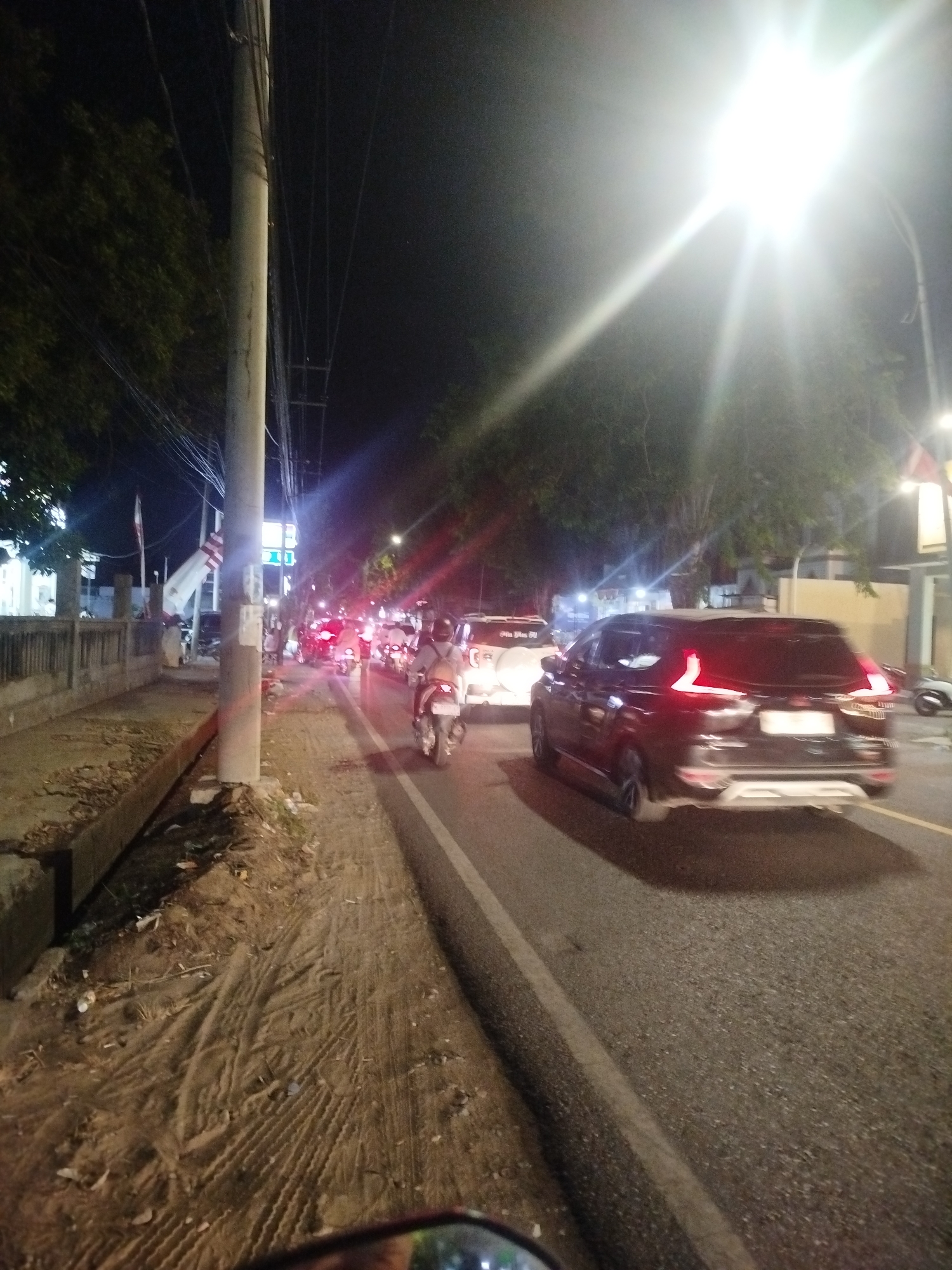 The image depicts a street scene at night in Burneh, Indonesia. A busy road is visible with several vehicles, including cars and motorcycles, in traffic. The lighting is dim, with the primary illumination coming from streetlights and the headlights and taillights of the vehicles. On the left side of the road, there is a sidewalk with a concrete barrier and some vegetation. The road surface appears to be asphalt, and there are lane markings visible. In the background, there are buildings and trees. The overall atmosphere is that of an active urban environment at night.
