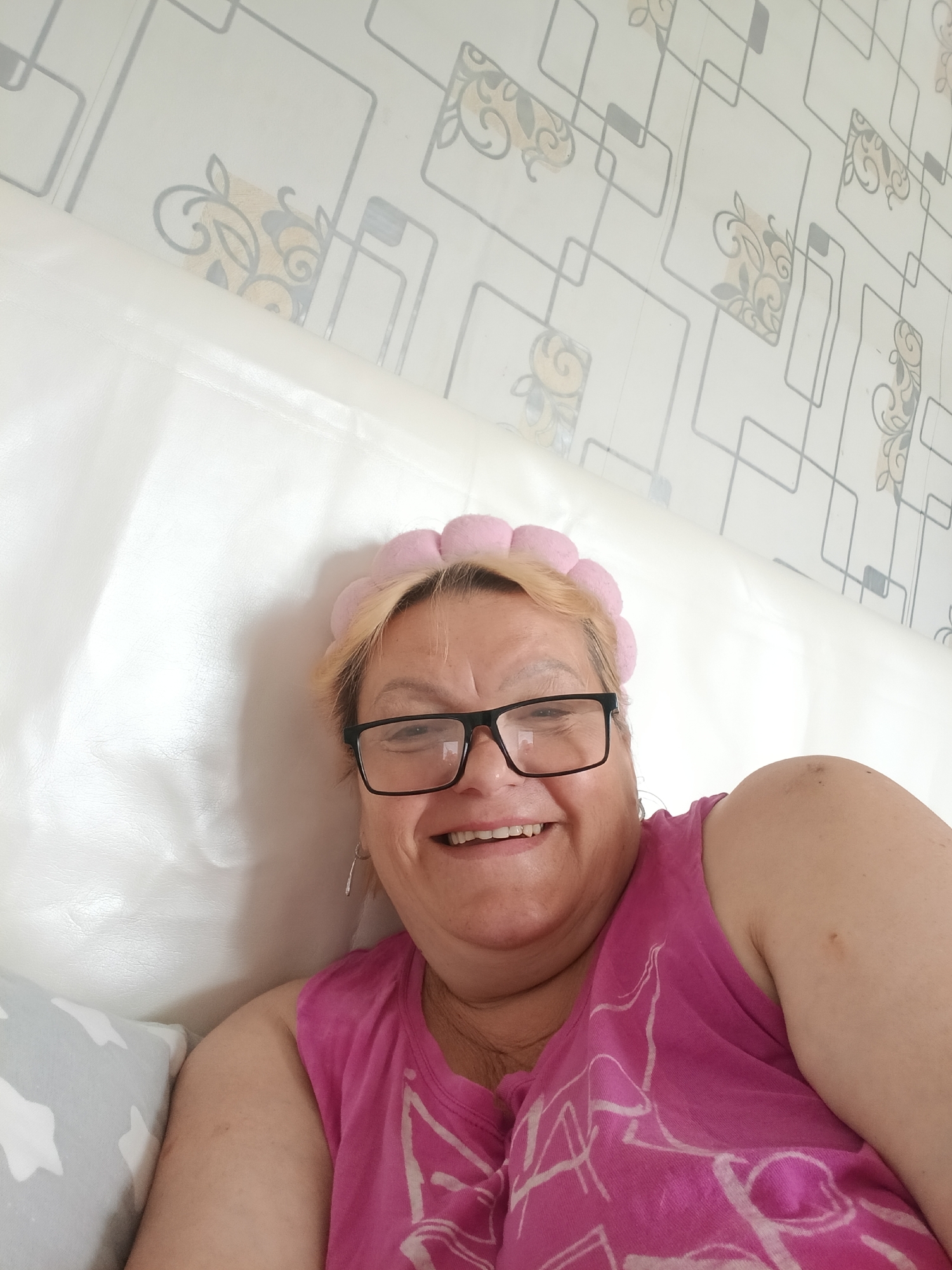 A woman with blonde hair and black framed glasses is lying down. She is wearing a pink shirt and a pink headband. The background includes a white headboard and wallpaper with a gray geometric pattern and beige floral designs. The location is Красный Маяк, Russia.