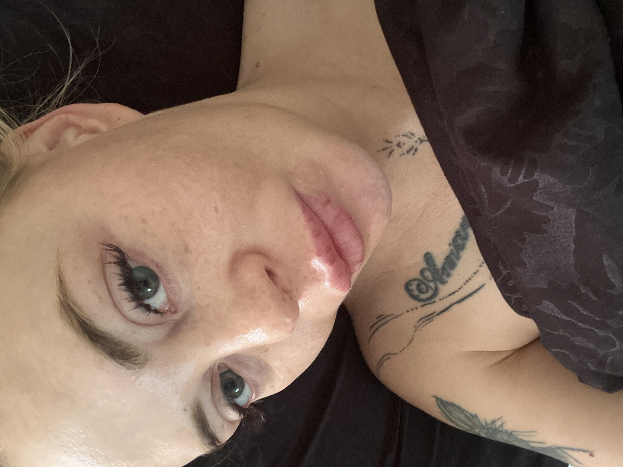 A close-up, overhead shot shows a young woman's face from the nose down, with one eye and part of her ear visible. She has light skin with freckles, green eyes with visible eyelashes, and full, pink lips that appear slightly chapped. Her eyebrows are dark and well-defined. She is partially covered by a dark brown fabric with a subtle patterned texture, which drapes over her shoulder and chest. A tattoo is visible on her left shoulder and upper arm, and another small tattoo is visible on her neck. Her hair is blonde and appears to be pulled back loosely. The lighting suggests it is daytime, possibly indoors, with soft shadows. The background is dark and out of focus.