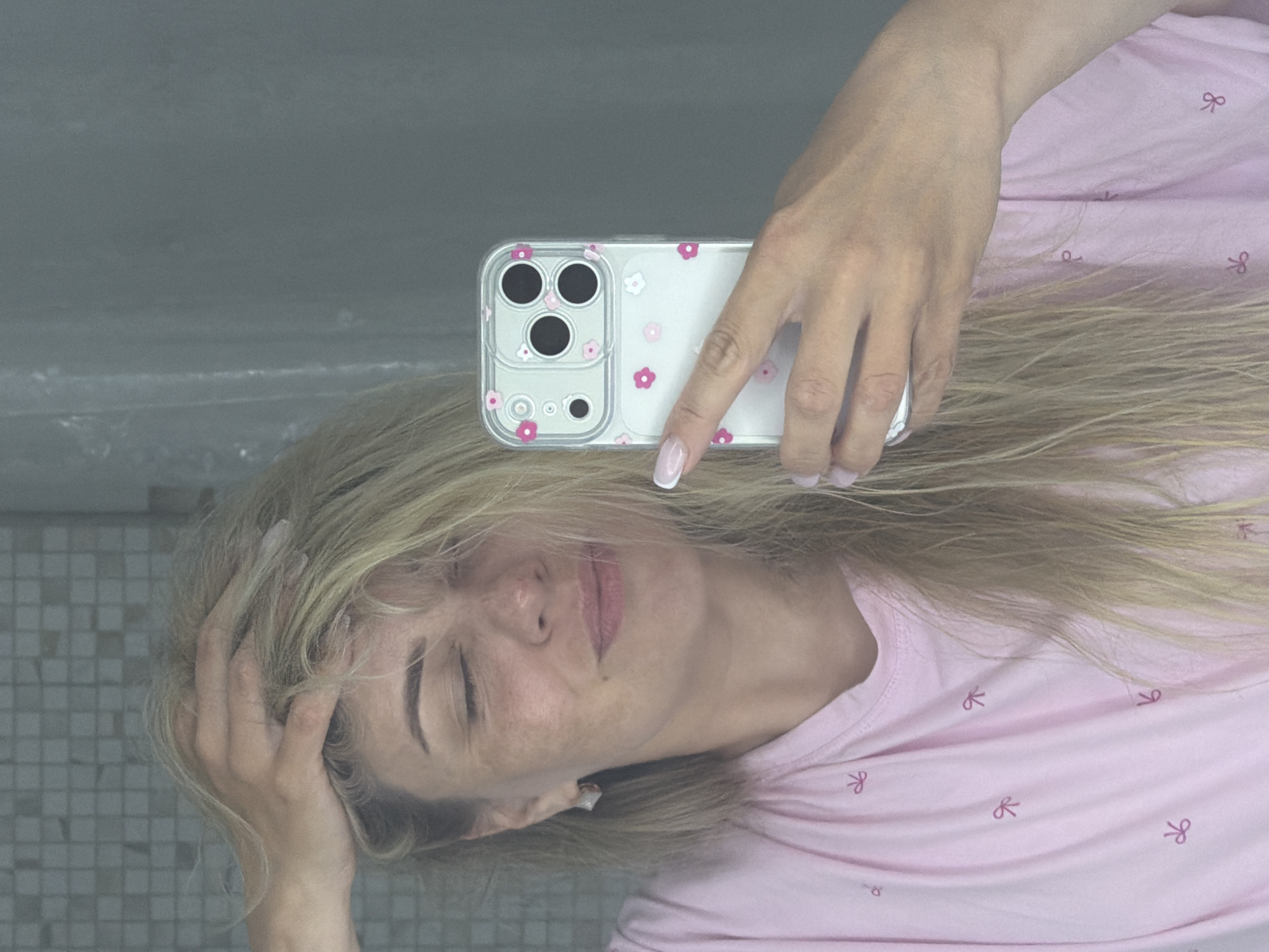 This media file captures a reflection of a woman taking a selfie with a smartphone. The woman has long, blonde hair spread around her head and is wearing a light pink t-shirt adorned with small, dark pink bow patterns. Her eyes are closed, conveying a sense of relaxation or contemplation, and her facial features are visible, including light freckles on her nose and cheeks. She appears to be holding the smartphone with her right hand (which appears as the left in the reflection), with her fingers curled around it. Her fingernails have a light pink and white French manicure.
The smartphone has a clear case decorated with small pink and white flower patterns. The camera module of the phone, featuring three lenses and a flash, is prominent.
The background of the reflection is split into two main areas: a darker, indistinct gray area occupying the upper part of the image, and a lighter gray area with small square tiles visible in the lower-left corner. The overall lighting is soft and diffused, not indicative of direct sunlight. No specific time of day can be determined, but the mood is calm. There is no visible text in the image. The location context provided is Votkinsk, Russia.