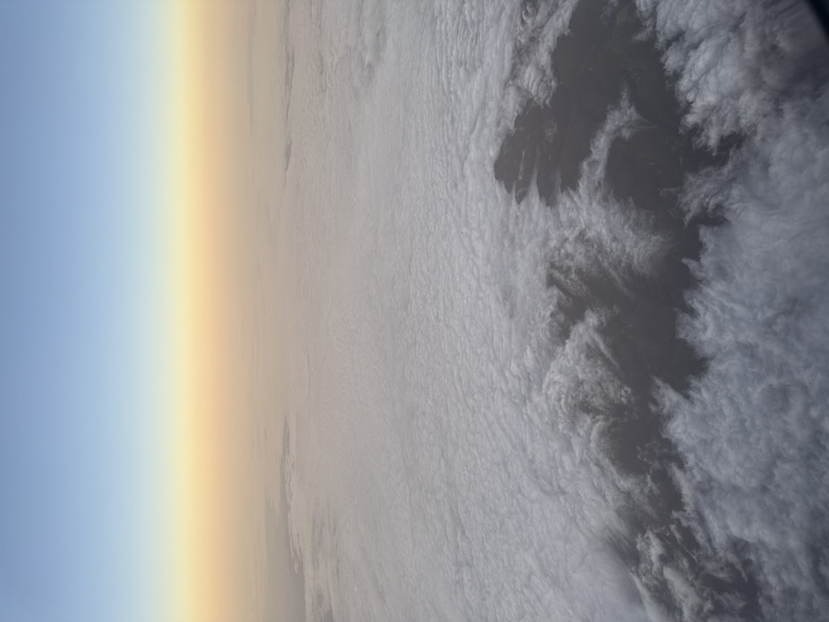 This aerial image depicts an expansive view of a cloud layer from a high altitude, potentially over the region of Santa María Xonacatepec, Mexico. The upper third of the frame shows a sky gradient, transitioning from a light blue at the zenith to a hazy, warm orange-yellow closer to the horizon line, indicative of either sunrise or sunset. The middle and lower sections are dominated by a dense, uneven blanket of white and light grey stratiform or cumulus clouds. Within this cloud cover, distinct, irregularly shaped dark grey to black landmasses are visible, breaking through or surrounded by the clouds, particularly in the lower-right quadrant and extending towards the center. These land features appear rugged, suggesting mountainous or hilly terrain partially obscured by the cloud layer. There are no discernible people, man-made structures, or specific actions visible within the frame. The scene presents a natural landscape from an elevated perspective, characterized by atmospheric haze and extensive cloud formations interacting with underlying topography.
