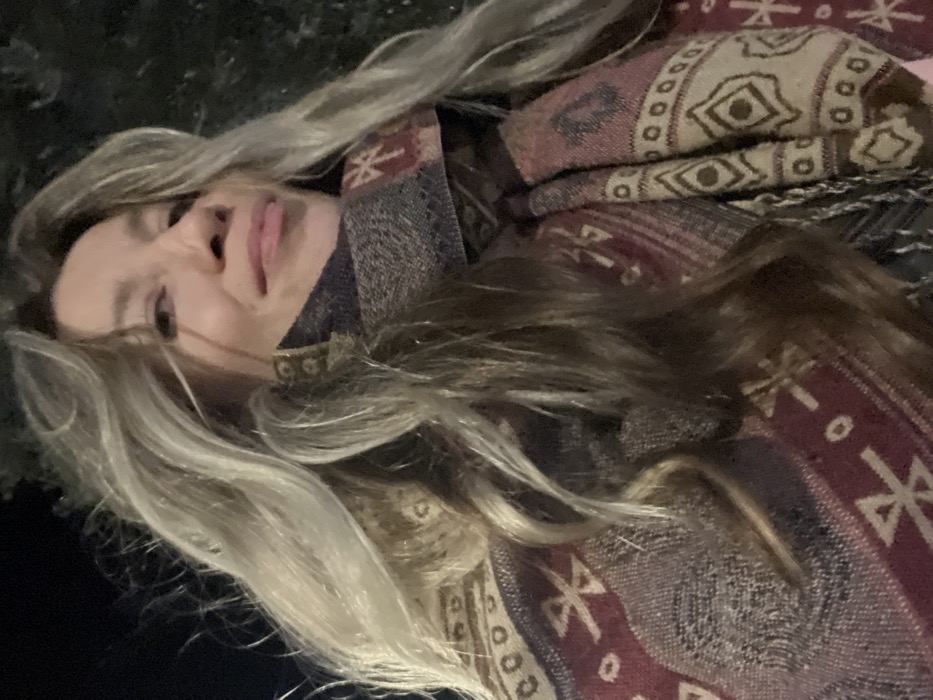 A young woman lies sideways, sticking out her tongue in a selfie. She has light blonde and brunette hair, and is wearing a patterned scarf or blanket with geometric and Aztec-like designs in brown, red, and beige hues. The background is dark, possibly outdoors. The photograph is shot at an angle, giving a casual and slightly playful feel.