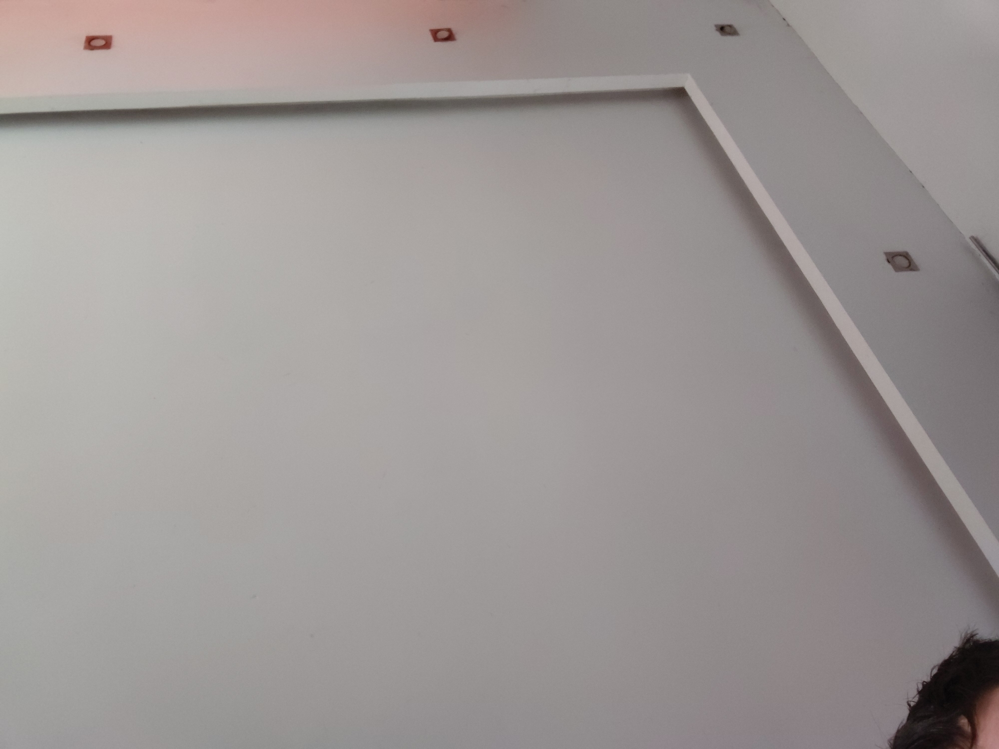 The image captures an indoor scene dominated by a light gray wall and ceiling. A prominent architectural feature consists of a large, raised rectangular border on the wall, creating a framed or recessed section. Several small, square-shaped cutouts, likely for recessed lighting fixtures, are visible along the top of this wall and extending onto the ceiling. More such cutouts appear on a diagonal architectural element on the right side of the image, which seems to be a raised beam or a corner detail. A thin, light-colored strip or rod runs parallel to this diagonal feature on the far right. The surfaces are painted in a uniform, light, neutral color. In the bottom right corner, the top of a person's head, featuring dark, wavy hair, is partially visible. No specific activity is depicted, and there is no discernible text. The setting suggests a modern and clean interior space.
