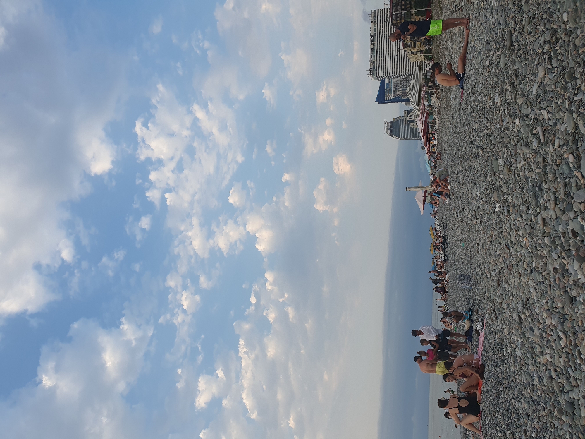 The image depicts a lively pebble beach scene in Batumi, Georgia. In the foreground, people are scattered across the beach, some sitting on towels, others standing or walking. The middle ground features more people, many relaxed on the shore, with umbrellas providing shade. In the background, modern high-rise buildings, characteristic of Batumi's urban landscape, are visible, including one with a distinctive rounded design and another resembling a striped tower. The sea is calm and stretches to the horizon under a sky filled with scattered white clouds. The overall atmosphere suggests a popular tourist destination on a pleasant day.
