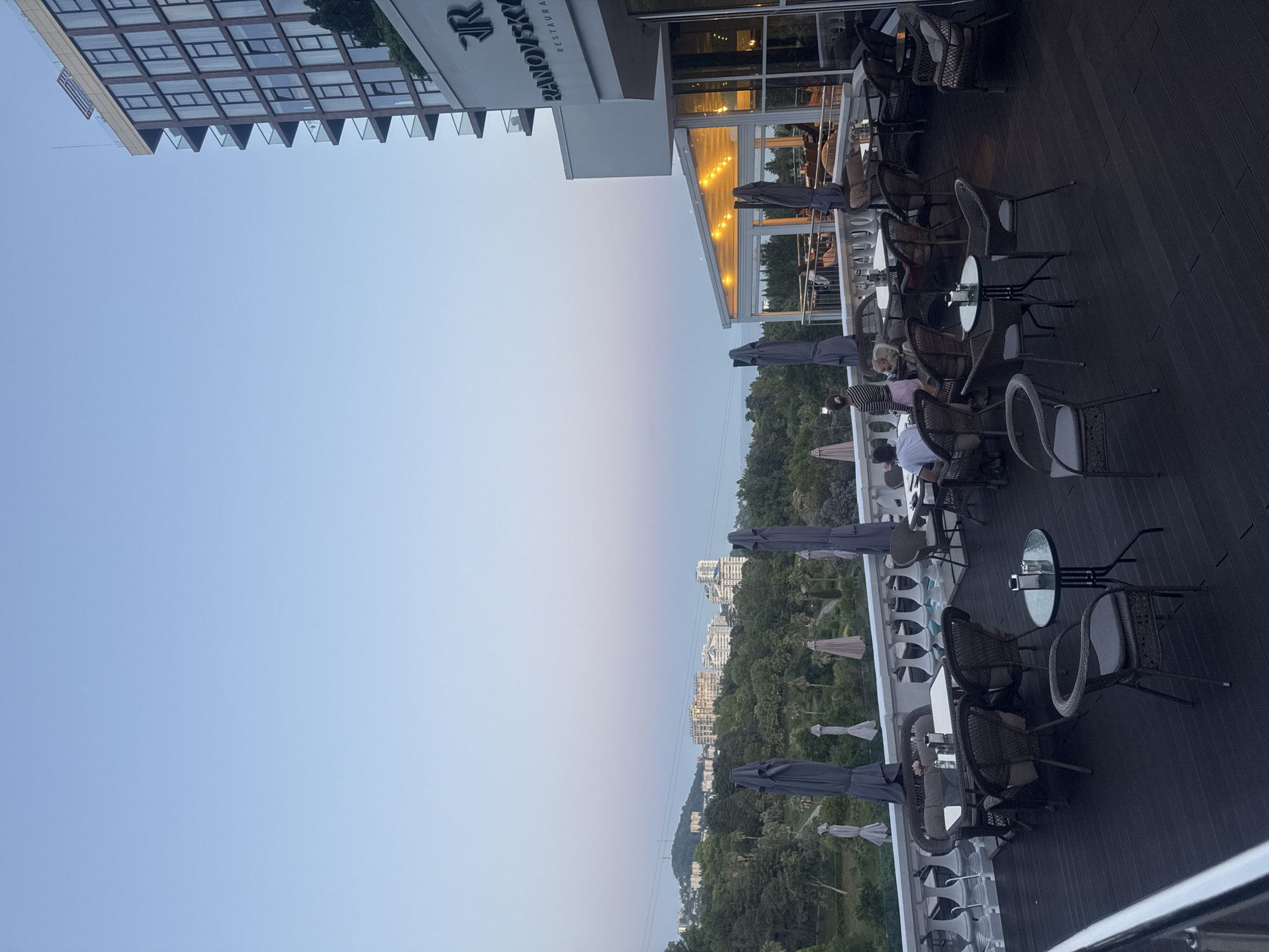 An exterior, low-angle, wide shot captures a terrace restaurant at dusk, overlooking a lush, green valley dotted with buildings and a distant sea. The sky is a pale gradient of blue and pink, indicating twilight. The terrace itself is made of dark wood planks and features multiple tables and chairs, some occupied by diners. Wicker chairs and tables with glass tops are arranged around the space. Several large, closed umbrellas are positioned along the railing. To the right, a modern, multi-story building with a facade of windows rises against the sky. A restaurant sign with the letter "R" prominently displayed is visible on the building. The background depicts a dense forest, rolling hills, and several mid-rise apartment buildings. In the far distance, a body of water can be seen. The scene is set in Sochi, Russia, specifically at the Ranovsky Restaurant.
