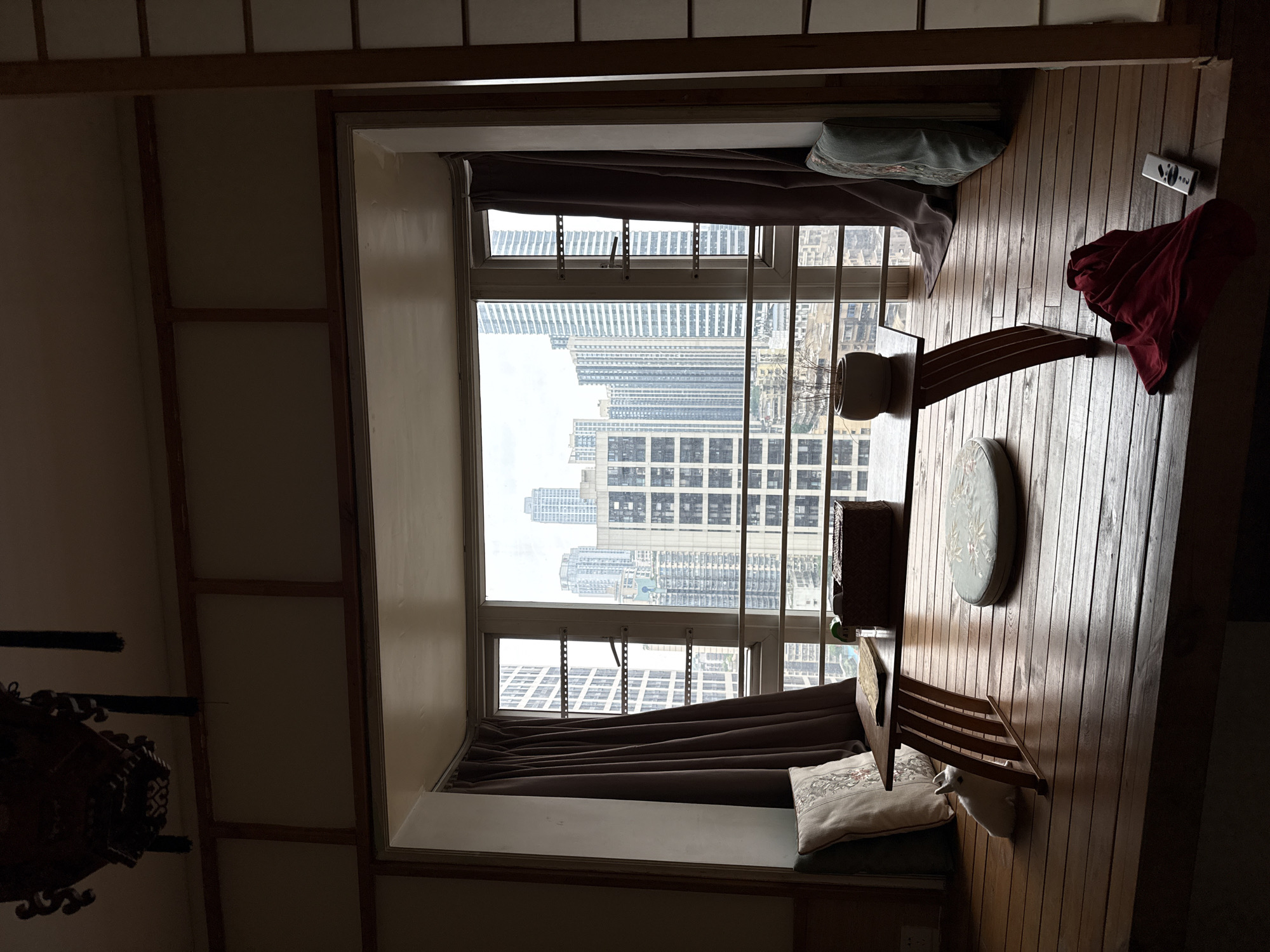 The interior scene depicts a room with a window framing a cityscape. The window is draped with brown curtains and security bars, providing a view of numerous high-rise buildings, characteristic of Yuzhong District, China. The room has wooden flooring and is furnished with a low table, cushions, and a folded chair. A red garment is draped over the edge of the table. A remote control lies on the floor. The room is illuminated by natural light filtering through the window.