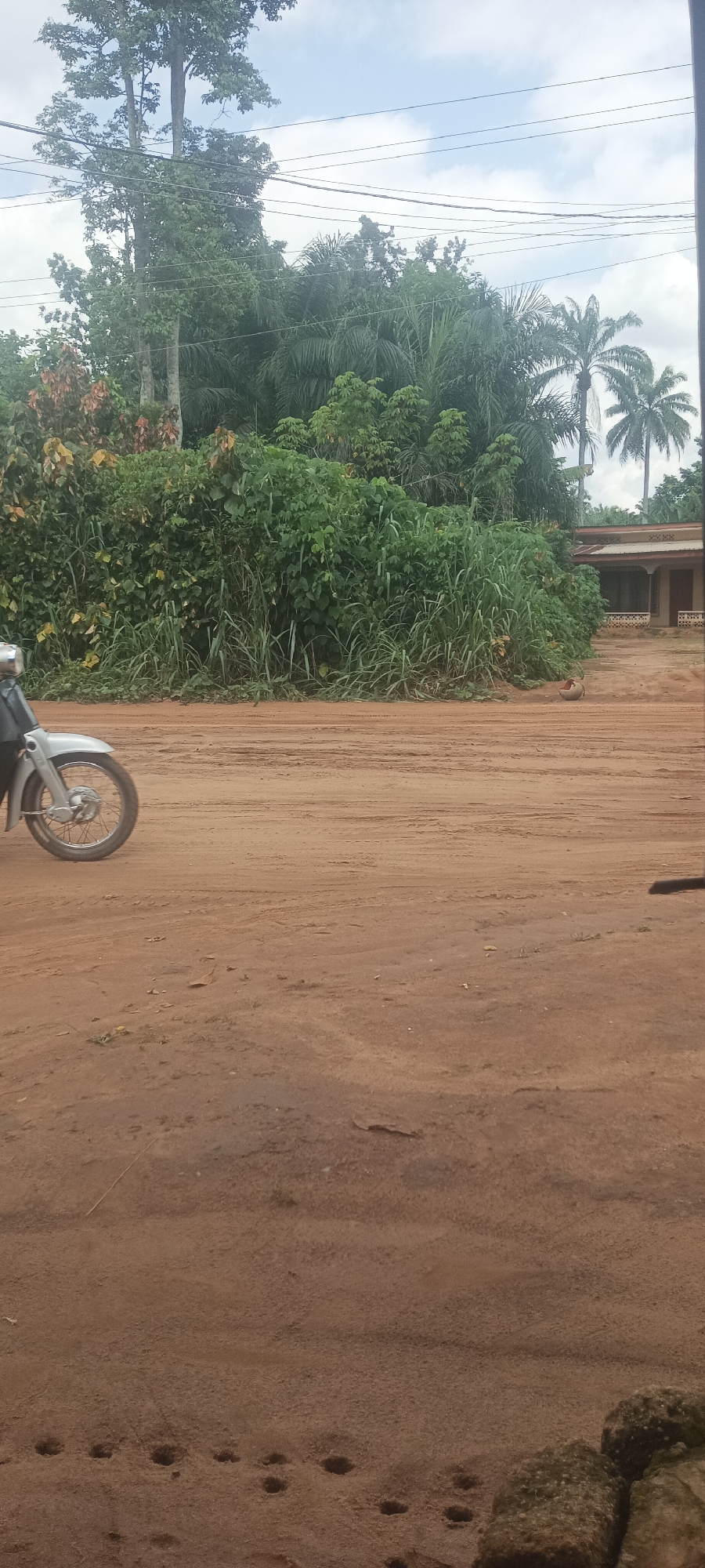The image shows a dirt road in Amauzu, Nigeria. A motorcycle is partially visible on the left side of the road, with its front wheel in view. Lush green vegetation lines the road. A building is visible in the background. The sky is overcast with scattered clouds. The road surface shows tire tracks and footprints.