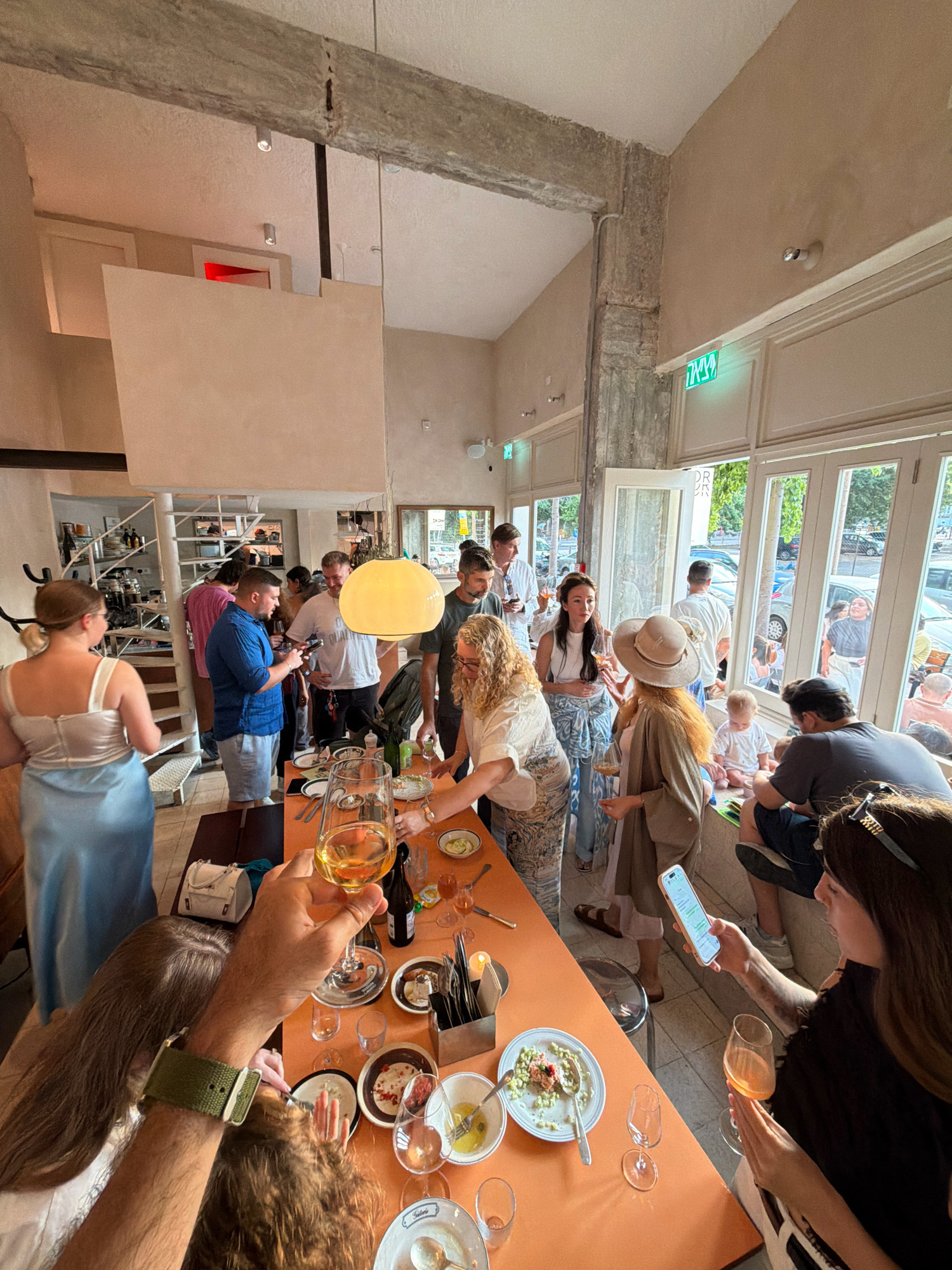 The image captures an indoor social gathering in Tel-Aviv, Israel, likely a restaurant or event space, characterized by natural light filtering through large windows. A group of people are seen mingling and seated around an orange table laden with food and drinks.
In the foreground, a person's arm, adorned with a green watch band, is holding up a glass of amber liquid. Another person in the foreground is looking at a smartphone, holding a glass of pinkish-orange liquid.
The environment features light-colored walls, exposed concrete beams, and a distinctive yellow pendant light fixture hanging from the ceiling. A spiral staircase is visible in the background, and a neon green exit sign is prominently displayed on the wall. Outside the windows, trees and cars suggest an urban setting.
People are dressed in casual attire, with several holding drinks and engaging in conversation. Some individuals are standing, while others are seated at the table. A young child is also present, seated on a ledge near the windows.
The overall scene depicts a lively and informal social event, possibly a meal or a pre-dinner gathering, in a modern and airy venue in Tel-Aviv.