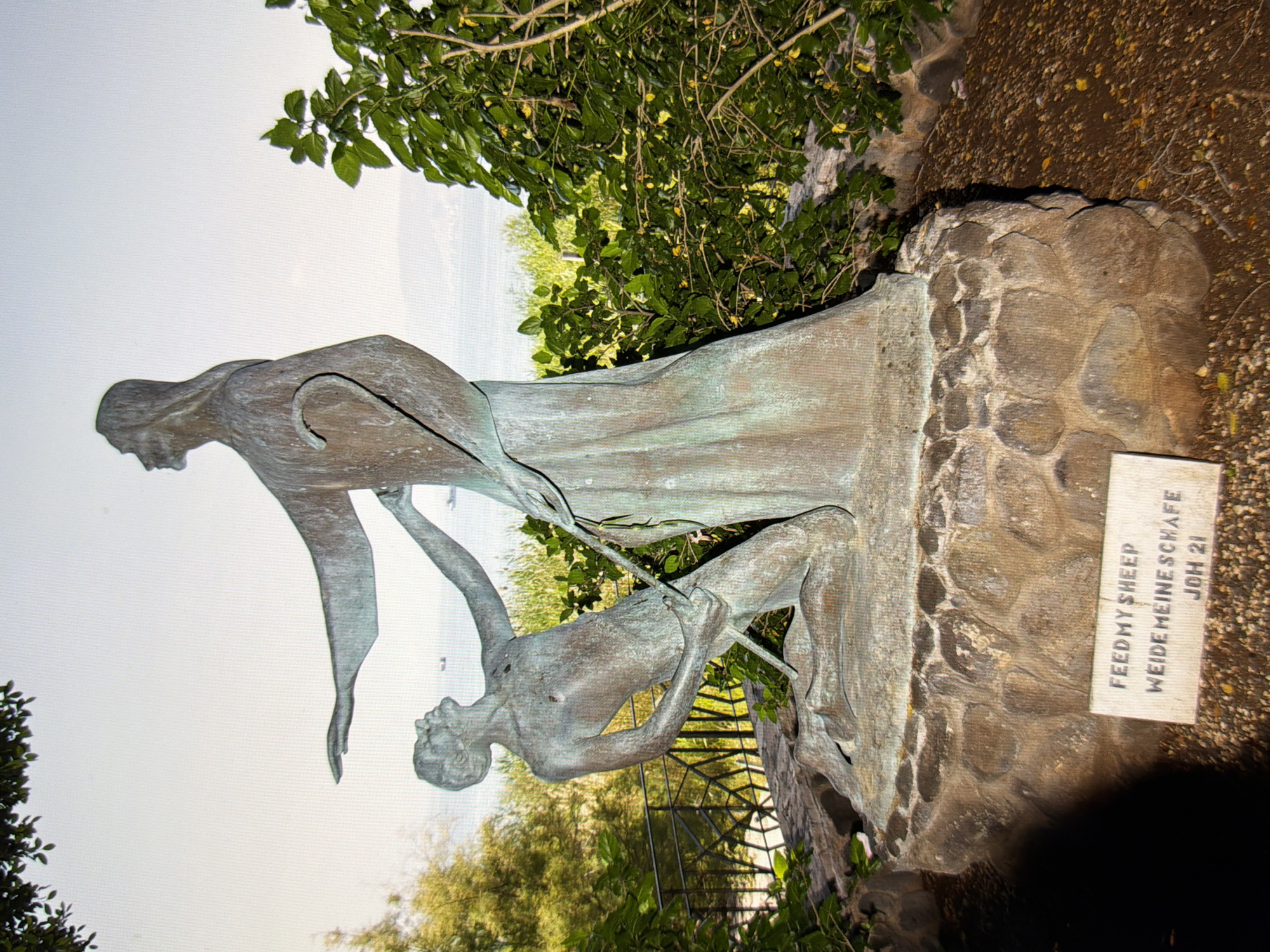 A bronze statue depicts Jesus Christ, robed and holding a shepherd's crook, standing over a kneeling man. The man looks up at Jesus with his arms outstretched, his right hand grasping the crook. The statue rests on a stone base, and a sign in front reads "FEED MY SHEEP WEIDE MEINE SCHAFE JOH 21." The background shows lush green foliage, trees, a body of water, and a hazy sky. The scene is set outdoors, likely in a park or garden. The statue's weathered patina suggests it is outdoors. The image does not provide any information about its location in Kyiv, Ukraine.