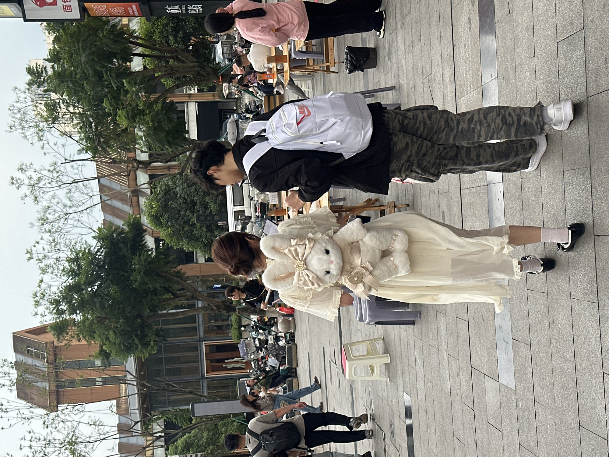 The image depicts a street scene in Chongqing High-tech Zone, China. A young woman in a light-colored dress with ruffles and a stuffed rabbit purse stands on a paved sidewalk. She wears pink socks and black shoes. Beside her, a person with a white backpack, wearing a black jacket and camouflage pants, stands facing her. In the background, other people are visible, some sitting at outdoor tables. Trees line the street. The overall environment appears to be a commercial or recreational area.