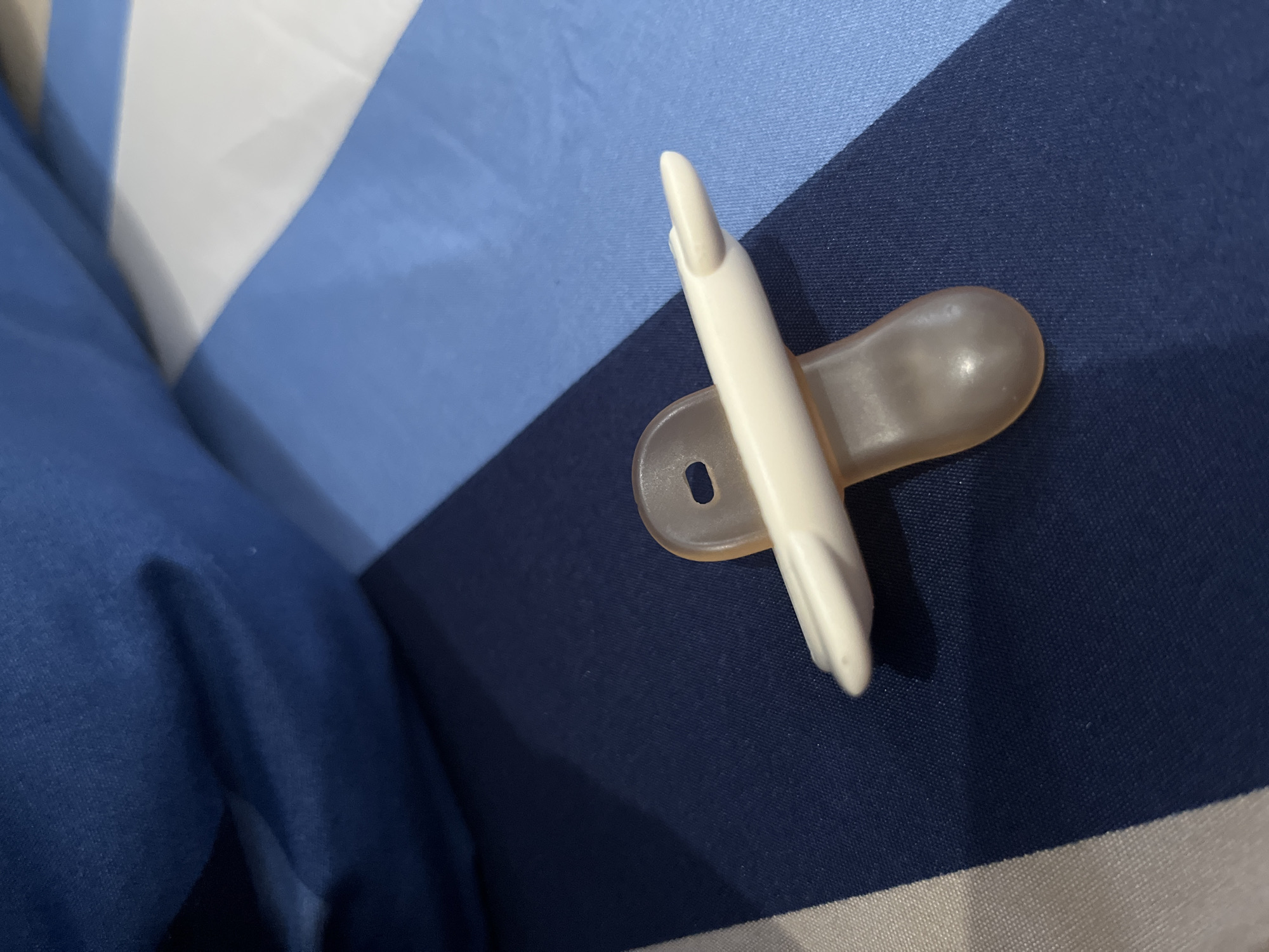 The image features a pacifier lying on a striped fabric surface. The pacifier is composed of two primary parts: a light-colored, possibly off-white, rigid plastic component and a translucent, amber-toned, soft silicone or rubber nipple. The plastic part acts as the shield and also incorporates a distinctive, elongated, and slightly curved projection, designed to mimic a finger or thumb for a baby's sucking comfort. A small, oblong hole is visible in the plastic shield, likely for ventilation or for attaching a pacifier clip.
The background is a fabric patterned with wide horizontal stripes in shades of blue and white. Dark navy blue, lighter blue, and white or very light gray stripes are visible, suggesting a bedsheet, blanket, or pillowcase. The fabric exhibits a subtle, tight weave. The lighting is soft and ambient, indicating an indoor setting, possibly with natural or artificial light; a slight shadow is cast by the pacifier on the darker blue stripe. There are no people present, and no specific activity is taking place other than the object resting on the fabric. No text is visible anywhere in the image. While the location context is Lhokseumawe, Indonesia, there are no specific visual cues within the image itself to denote this geographic location.