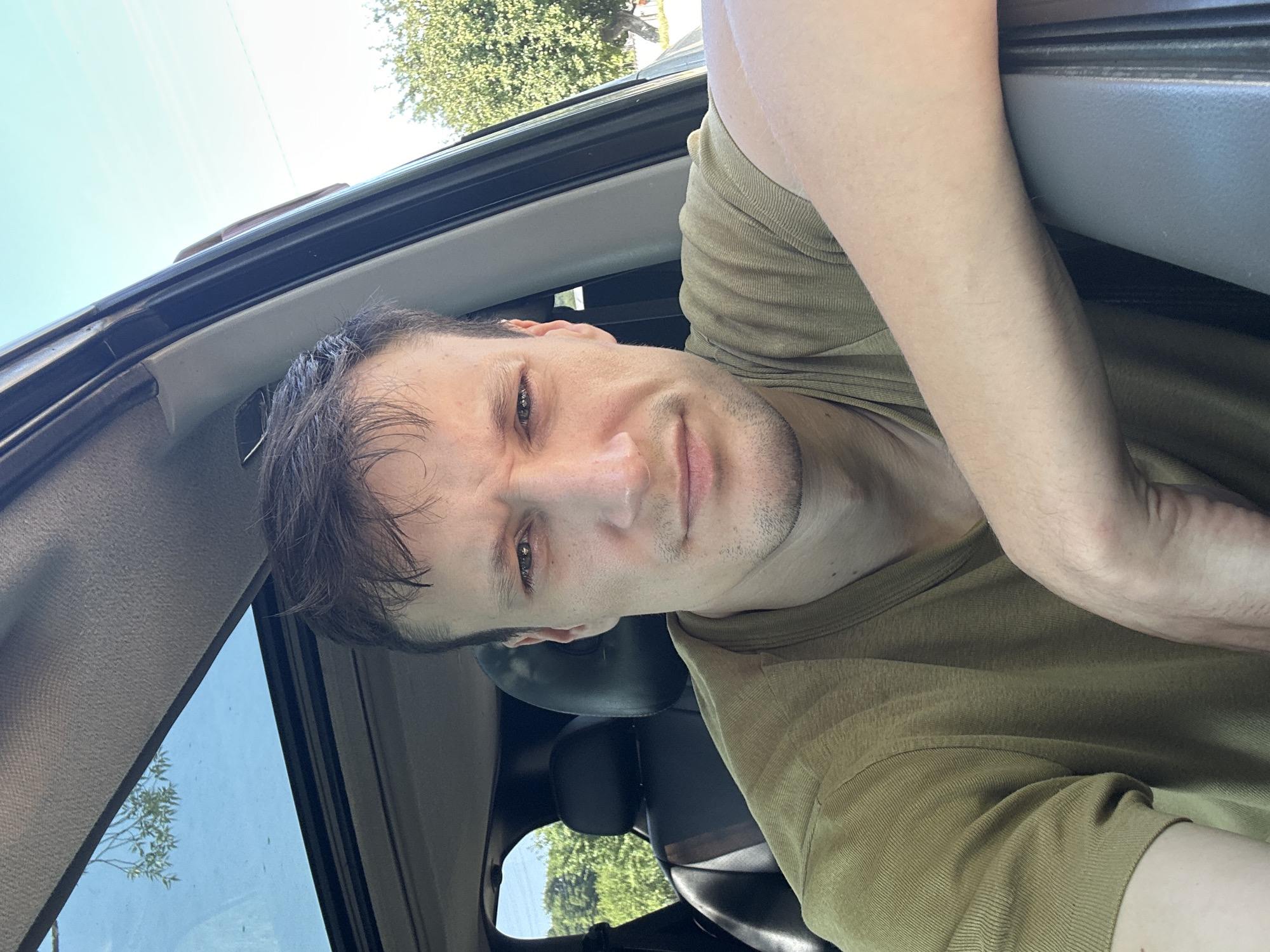 A selfie taken from inside a vehicle shows a man in his late 20s or early 30s with short, dark brown hair. He has a light complexion and is wearing a green t-shirt. The man is leaning against the open car door, with his right arm resting on the door frame. His left arm is visible in the foreground, bent at the elbow. The interior of the vehicle shows black leather seats and a sunroof. Outside the car, a green, leafy tree is visible against a clear blue sky. The scene appears to be set in a rural or suburban area, likely Makiv, Ukraine, given the context. The lighting suggests it is daytime, possibly late morning or early afternoon.