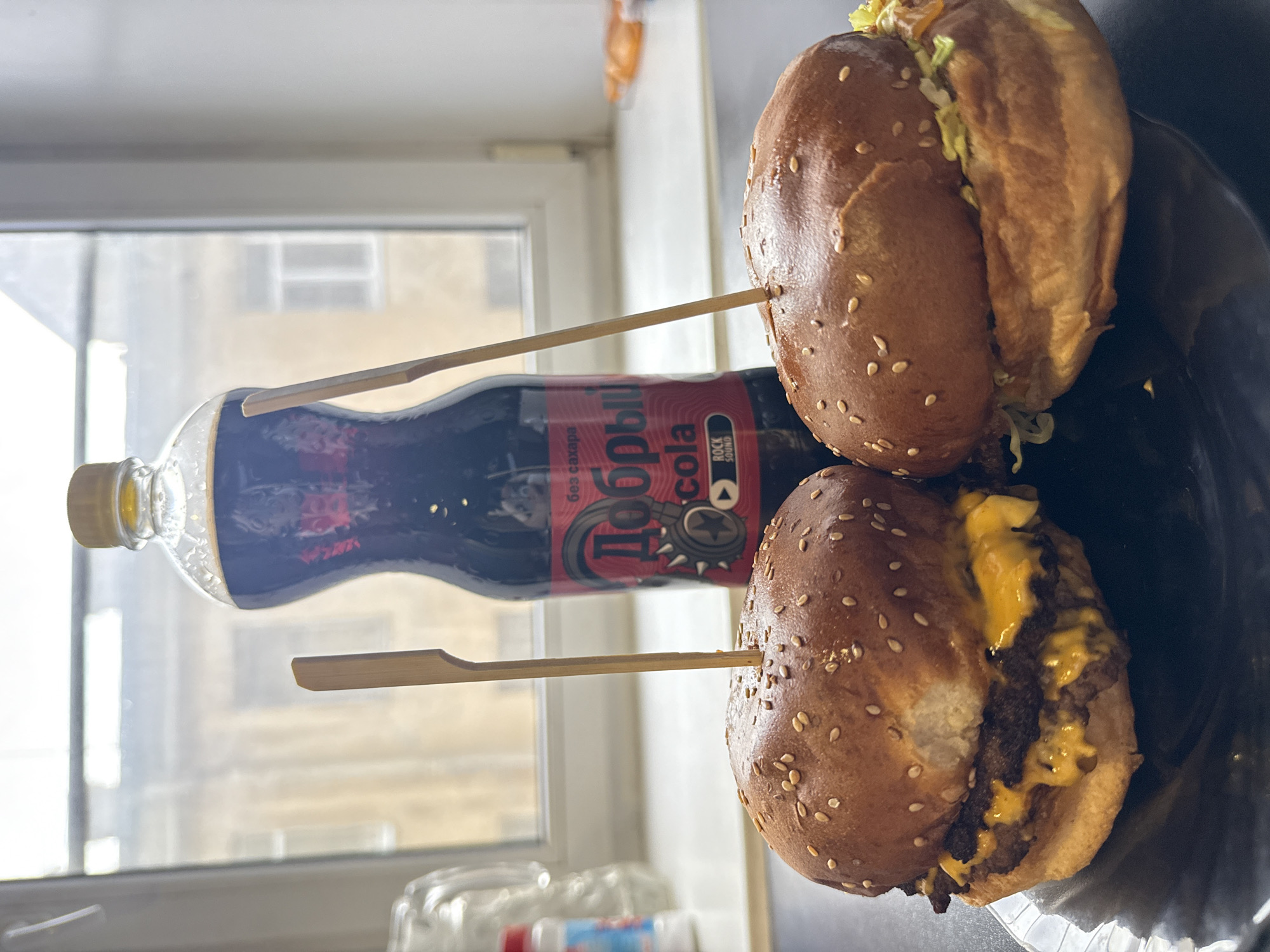 The image displays a meal consisting of two burgers and a bottle of soda, arranged indoors next to a window. The overall scene is brightly lit by natural light, indicating it is daytime.
Two burgers, each with a sesame seed bun, are visible, lying on their sides on a dark, possibly black, plate or container. The lower burger prominently features melted orange cheese over two meat patties. The upper burger shows visible green lettuce as a topping. Both burgers are pierced by wooden skewers, which appear to extend from the soda bottle.
A clear plastic bottle containing a dark-colored cola beverage stands vertically between the burgers. The bottle has a yellow cap. Its label features the prominent text "Добрыи́ Cola" (Dobriy Cola) in red and white, along with a stylized star and gear emblem. Below this, the phrase "БЕЗ САХАРА" (BEZ SAKHARA), meaning "sugar-free," is visible, as is a small black box containing "ROCK SOUND."
The background shows a large window with a light-colored frame. Through the window, an out-of-focus exterior view of a building with multiple windows can be seen, suggesting an urban environment, consistent with the Saint Petersburg, Russia context. The arrangement of the food suggests a casual meal, possibly takeout.