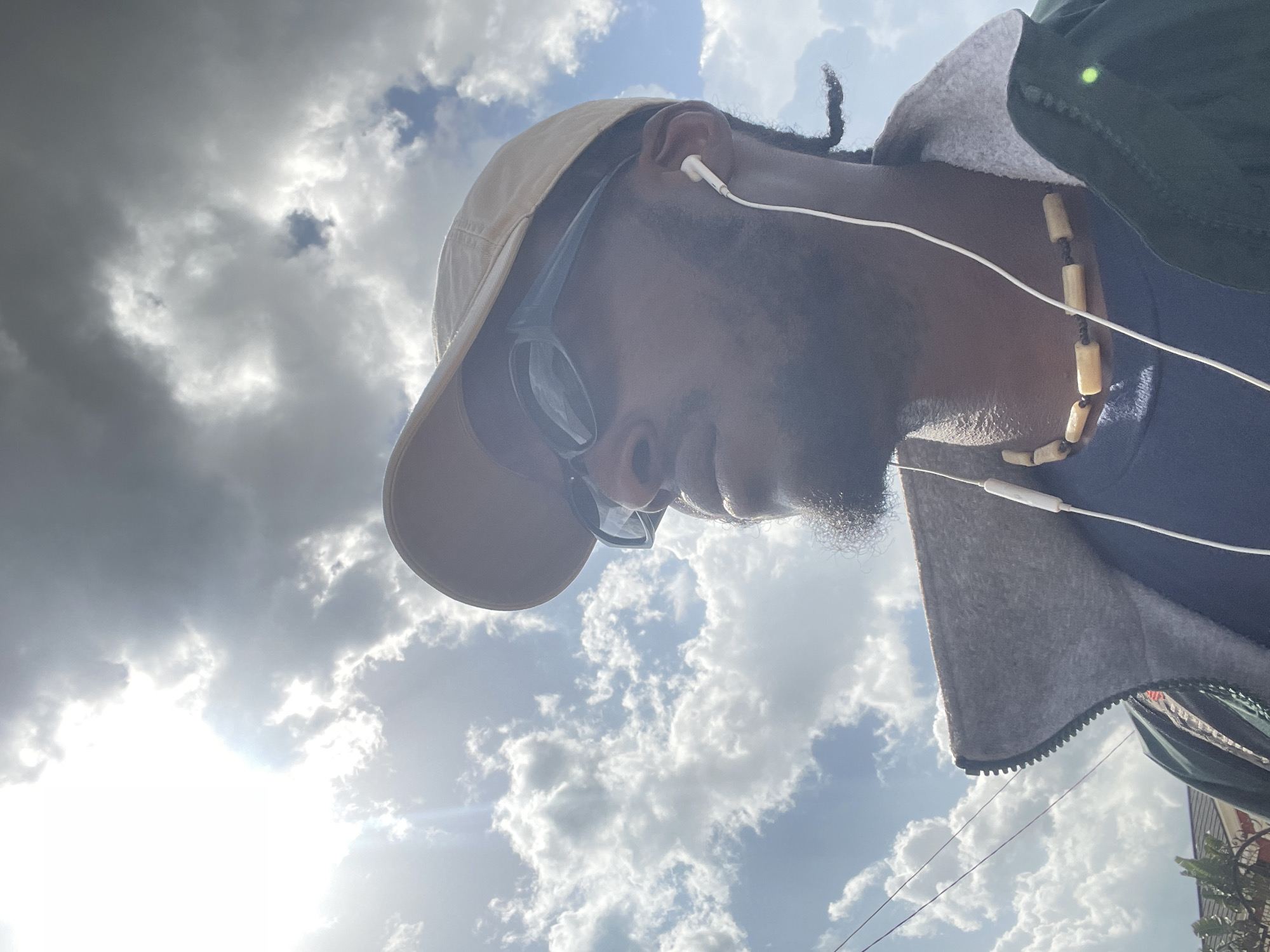 In a low-angle shot, a Black man wearing a tan baseball cap, sunglasses, a dark t-shirt, a green jacket with a gray collar, and a beaded necklace is seen from the chest up, looking to the left. White earbuds are plugged into his ears, with the cord extending down. The background is dominated by a cloudy sky with bright sunlight filtering through. The scene appears to be outdoors in the city of Jos, Nigeria, with a hint of residential buildings and foliage visible in the lower left corner. The overall impression is of a casual outdoor moment.