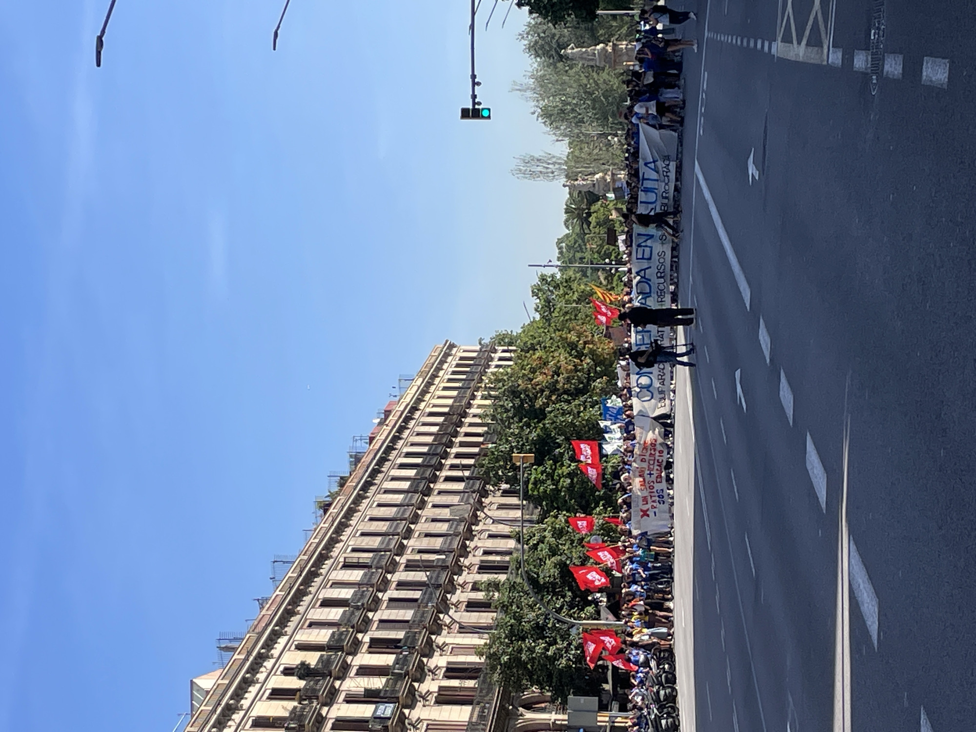 A protest or demonstration is occurring on a city street. The street is lined by a tall building. Numerous people are gathered, carrying flags (predominantly red) and banners. One banner reads (translated from the original Spanish): "NO MORE SALARIES, RATIOS + RESOURCES, SOS EDUCATION." Another banner appears at the front of the group, reading (translated from the original Spanish): "CONCERTED IN FIGHT, EQUALITY, RATING RESOURCES, BUREAUCRACY." A green traffic light is visible in the background.
The location is Barcelona, Spain.