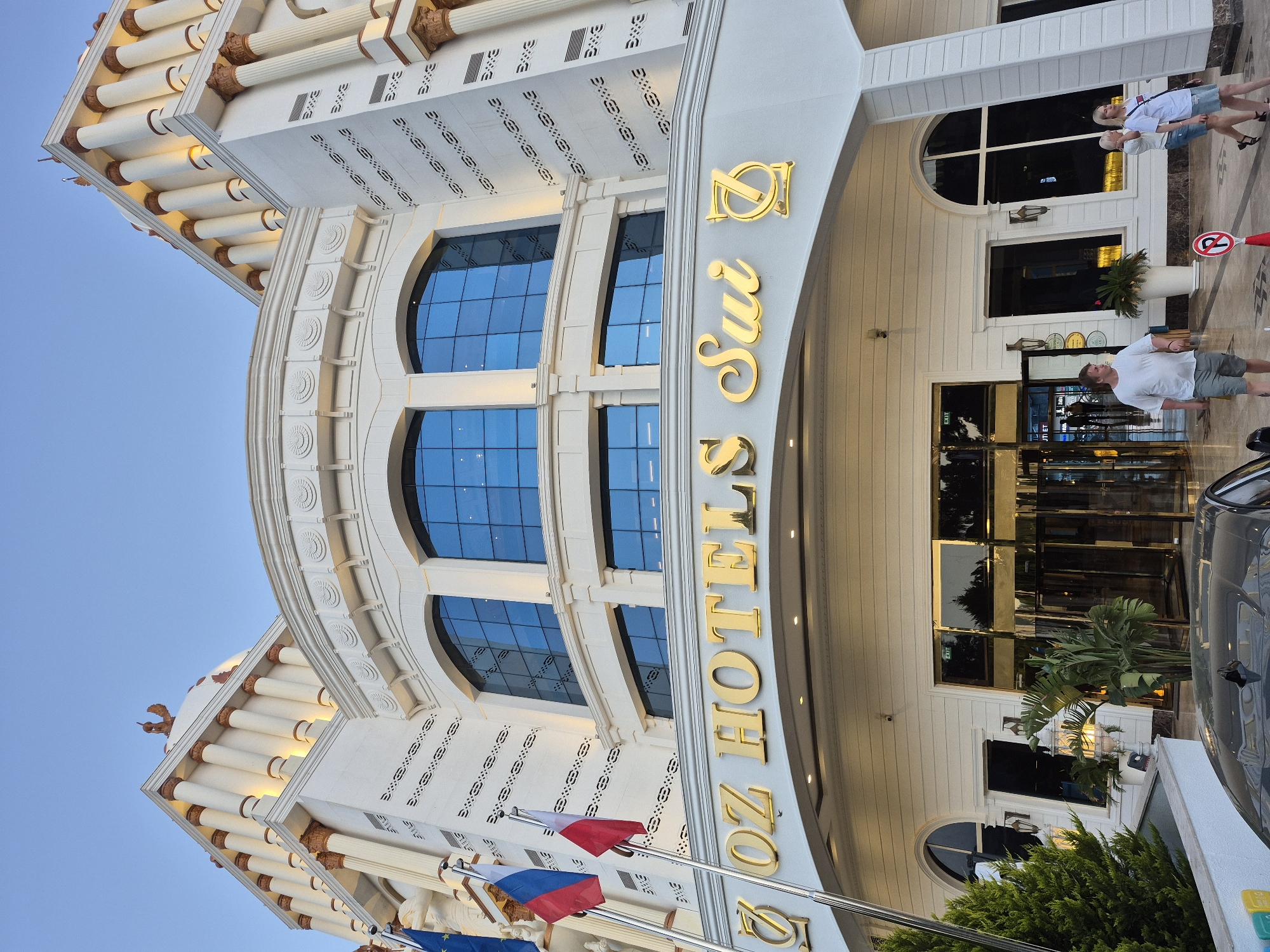 The image depicts the exterior of the Oz Hotels Sui, a hotel located in Alanya, Turkey. The building's facade is white with architectural details, including pillars, arched windows with blue glass, and decorative moldings. Gold lettering spelling out "Oz Hotels Sui" is prominently displayed above the entrance. Several flags are visible in the foreground. People can be seen entering and exiting the hotel. A car is parked in front. The sky is a clear blue.