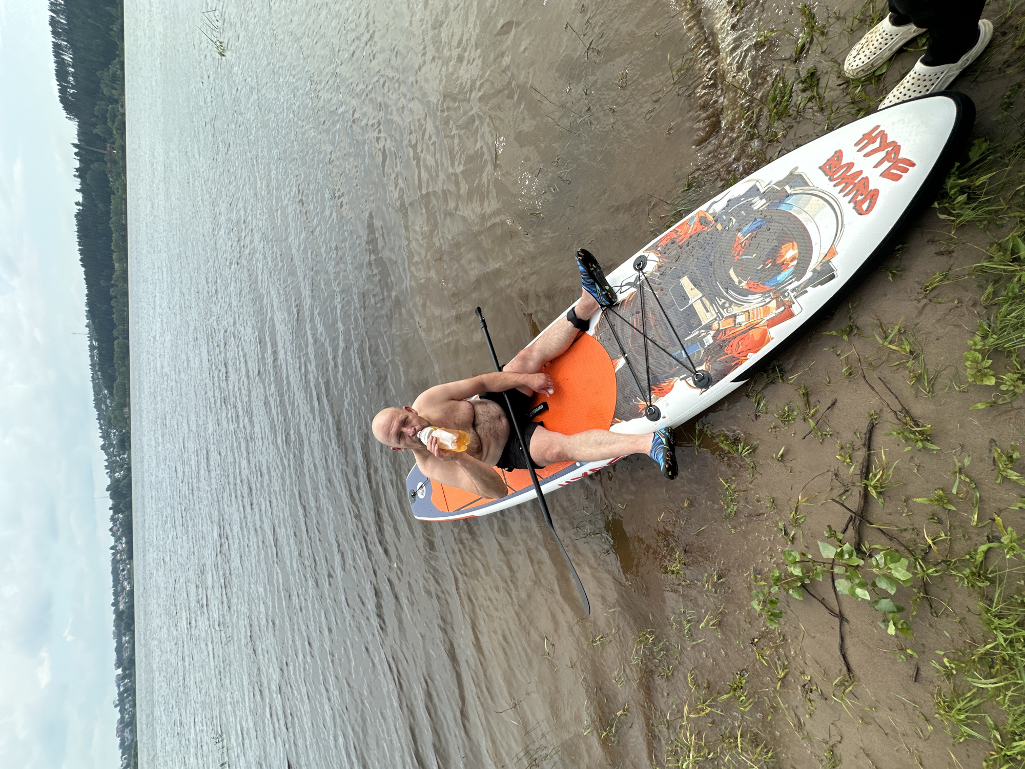 A bald, shirtless man is sitting on a white and orange stand-up paddleboard in shallow water near the shore of a large body of water. He is drinking from a yellow bottle. The paddleboard has "HYPE BOARD" written on its front in red lettering and a graphic design featuring what appears to be a camera. He is wearing black shorts and blue and black water shoes. The shore is muddy with some green grass and small plants. In the background, across the water, is a dense line of trees and a small settlement with houses. The sky is overcast and cloudy. The location is the city of Noviy, Russia.