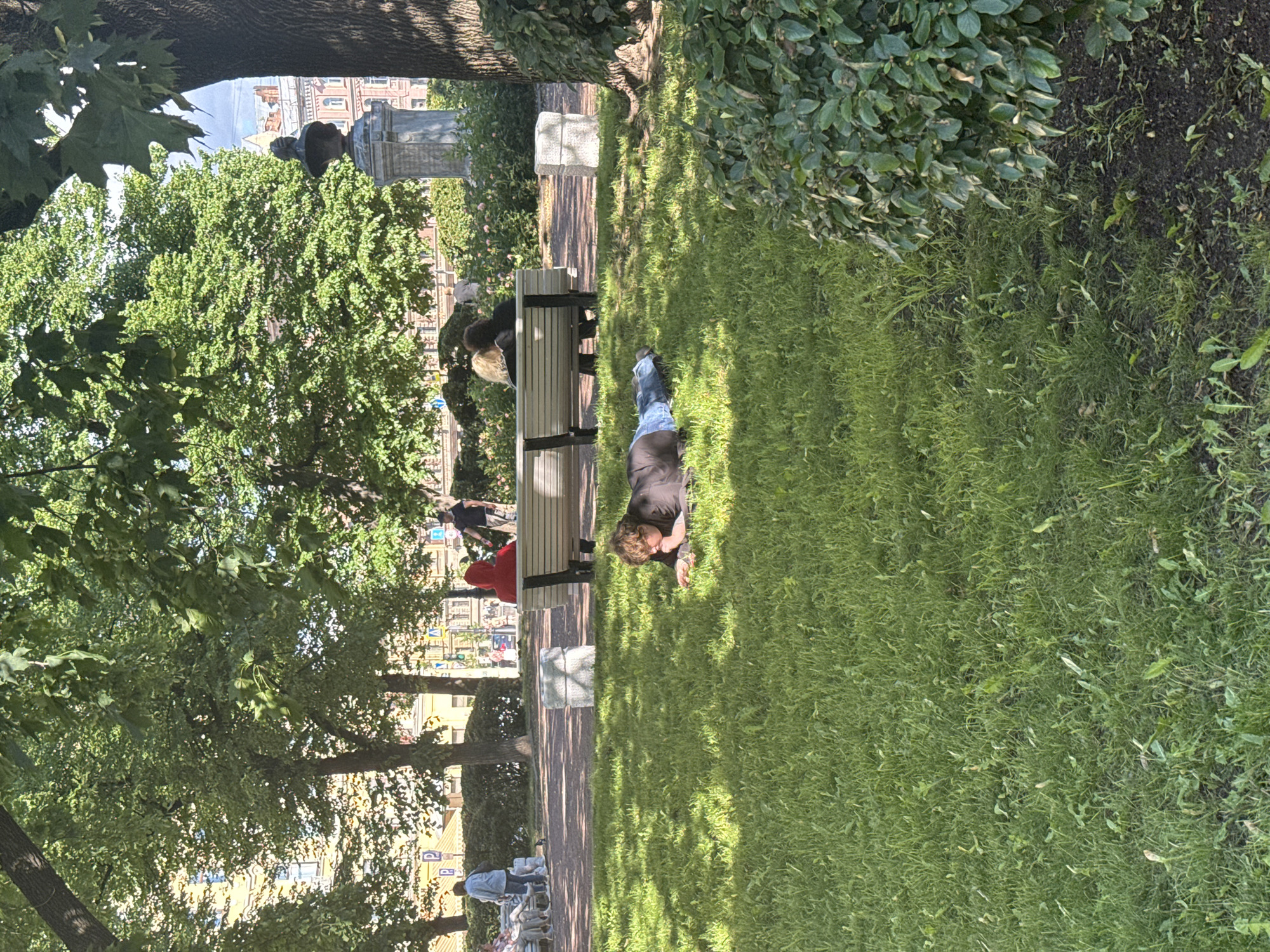 A man lies on the grass in a park, his head propped up on his hand. He is wearing a black t-shirt and blue jeans. Sunlight casts dappled shadows on the grass from the surrounding trees. In the background, a park bench is occupied by other people, and a pathway leads further into the park. Beyond the trees, buildings with red brick facades are visible, suggesting an urban setting within the park. The time appears to be daytime, with bright sunlight indicating fair weather. The overall scene suggests a relaxed atmosphere in a city park, possibly in Saint Petersburg, Russia, given the architectural style of the buildings. No visible text is present in the image.