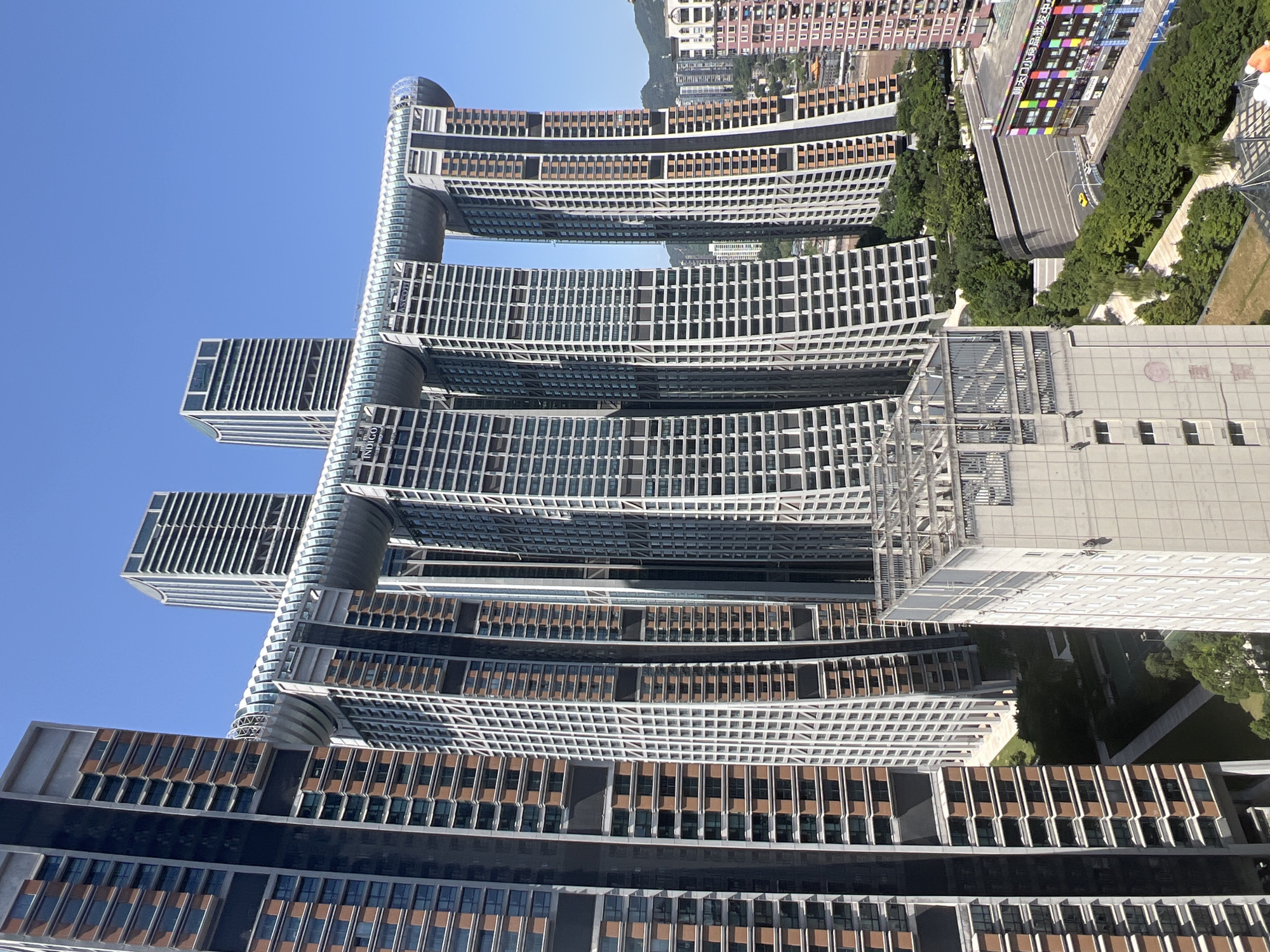 The image shows the elegant and futuristic "Crystal" structure of the Raffles City Chongqing in Yuzhong District, China. This iconic skyscraper complex features multiple towers connected by a skybridge, resembling a ship sailing through the sky. The photograph captures the towers from a slightly elevated perspective, showcasing their unique architectural design, with a clear blue sky as the backdrop. The surrounding urban landscape includes other buildings, greenery, and a hint of mountainous terrain in the distance. The overall scene is one of modern urban development and architectural innovation.