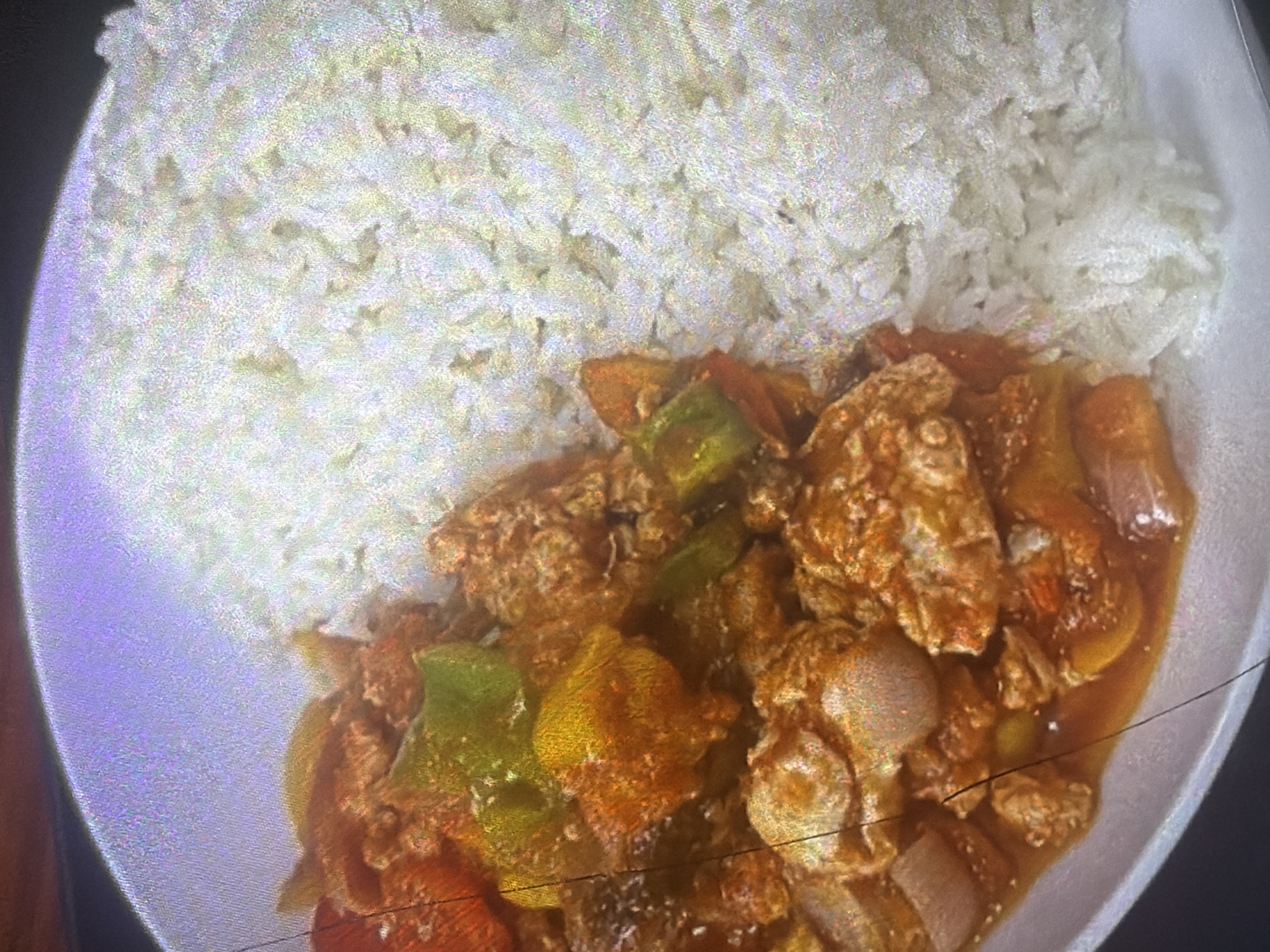 The image shows a close-up, overhead view of a white plate containing a meal. The plate is divided roughly in half. On the left side, there is a saucy dish with chunks of meat, bell peppers (green, yellow, and red), and onions in a reddish-brown sauce. The right side of the plate is filled with white rice. The overall lighting is somewhat dim and grainy, suggesting it might be taken indoors. There are no people visible in the image. The location is not specified in the image or the prompt, therefore it cannot be included in the description.