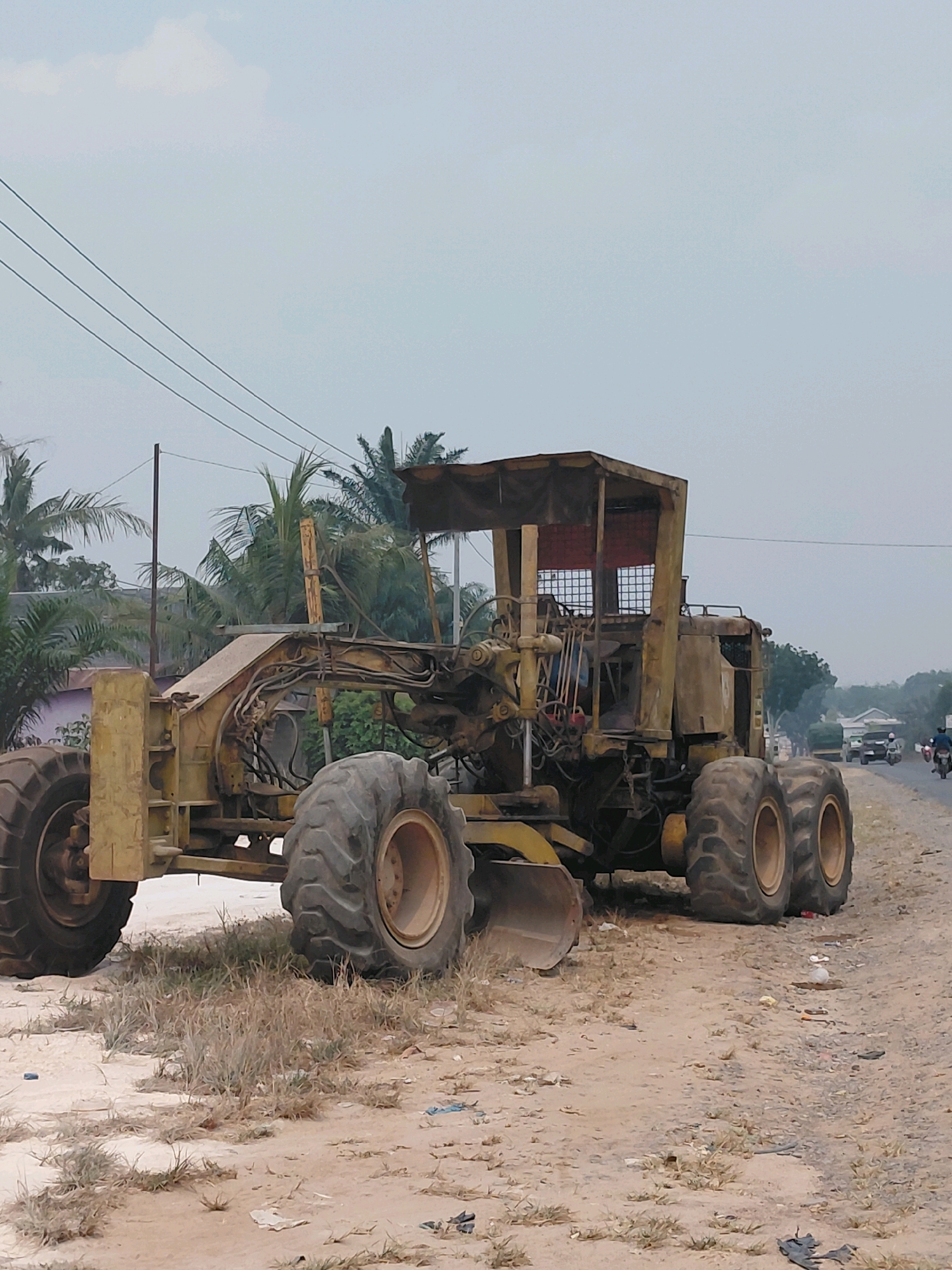 A damaged yellow grader is parked on the side of a dirt road in Persiapan Mulya Raya, Indonesia. The grader appears to be abandoned, with rust and debris visible on its frame and tires. Palm trees and other vegetation surround the area, and power lines stretch across the sky. In the background, a few vehicles, including cars and motorcycles, can be seen driving on a paved road. The overall scene suggests a rural or semi-urban environment that may be undergoing construction or experiencing a lack of maintenance.