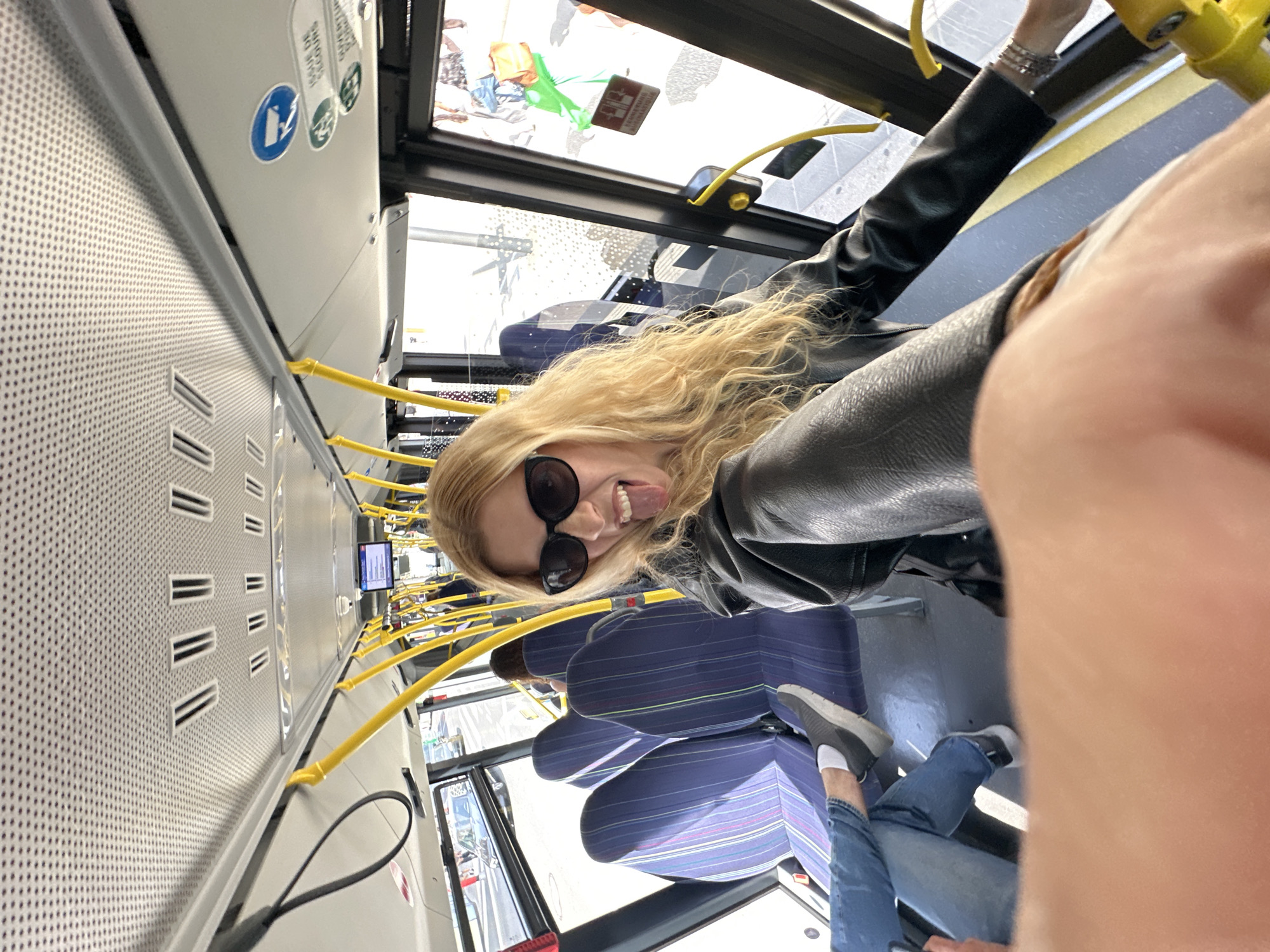 A woman with blonde hair and sunglasses is taking a selfie inside a bus. She is sticking her tongue out at the camera. The bus has blue seats with colorful stripes and yellow poles. Outside the bus, a street with cars and buildings can be seen. The scene appears to be in Luxembourg City, Luxembourg, given the context provided by the user's prompt.