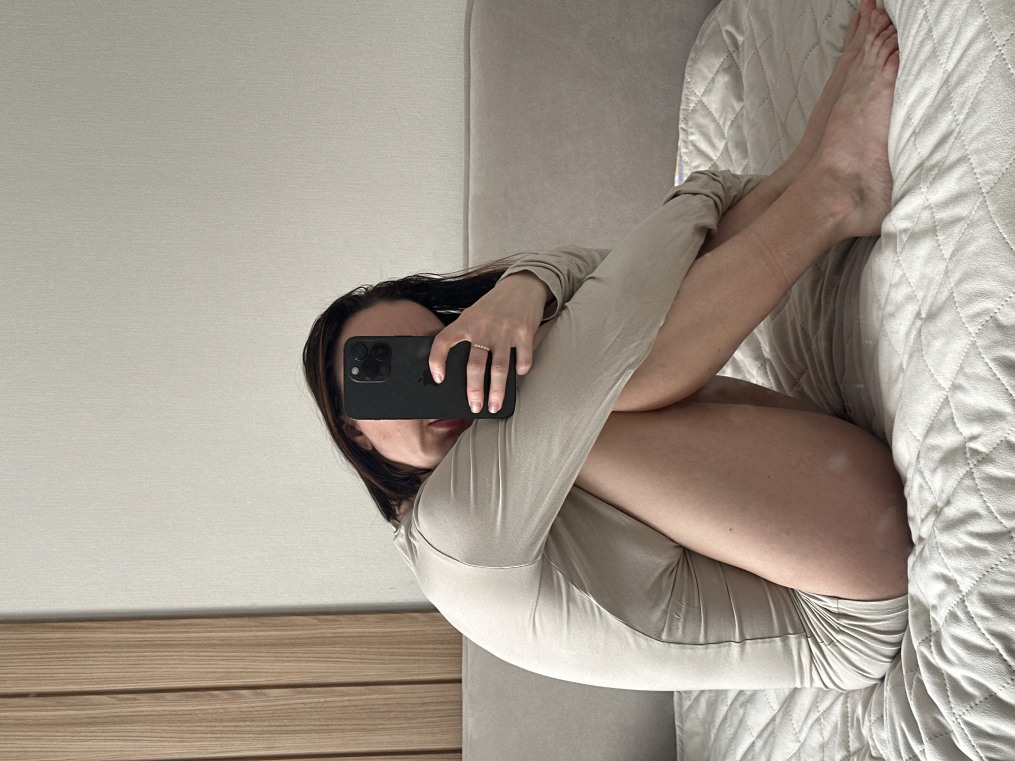 A single individual is depicted seated on a bed indoors in Votkinsk, Russia. The subject, a female with dark hair, is positioned with her knees drawn towards her chest, embracing her legs. She wears a form-fitting, long-sleeved, light beige garment. Her right hand holds a dark-colored smartphone, featuring multiple camera lenses, which partially obscures her face, indicating the action of taking a selfie. A ring is visible on the ring finger of her right hand. Her lower legs and feet are bare. The bed is covered with a light-colored, quilted blanket. Behind the individual, a light grey upholstered headboard is present, backed by a plain, light-colored wall. To the left of the frame, a section of vertical light-colored wooden paneling is visible, suggesting a decorative wall element.