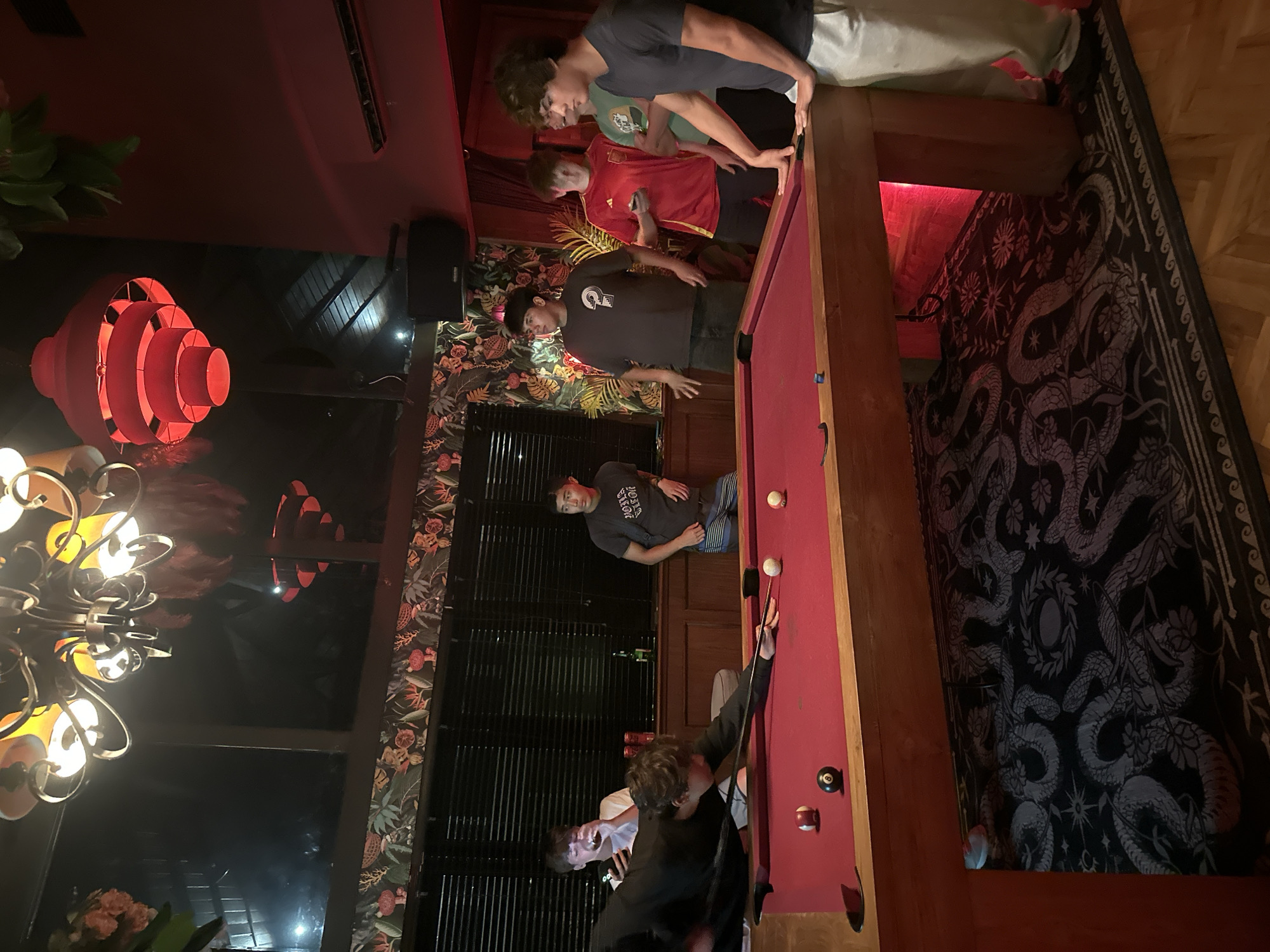 The image depicts an indoor scene with several young men gathered around a red pool table. The room features dark walls adorned with a patterned wallpaper and a large window with blinds. A chandelier hangs from the ceiling, and a red lantern-style light fixture is also visible. The floor is covered with a dark carpet featuring a dragon-like pattern, and wooden parquet flooring is visible around the edges.
In the foreground, a young man in a black shirt is actively playing pool, leaning over the table with a cue stick, preparing to strike a white ball. Another young man in a grey t-shirt and striped shorts stands to the right, observing the game. Further back, several other young men are visible: one in a white t-shirt is holding a phone and a drink, another in a grey t-shirt with a question mark graphic is looking towards the table, and one in a red shirt is also looking in the direction of the pool game. The atmosphere appears to be casual and social.
The specific location is a residential property in North Kuta, Indonesia.