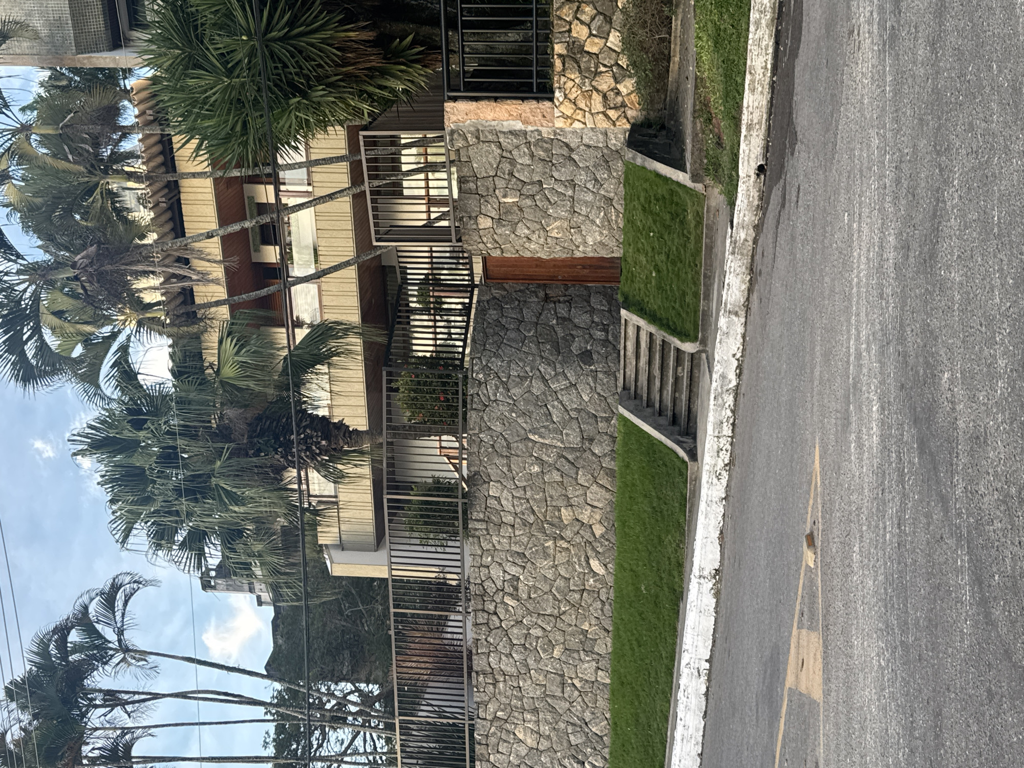 The image depicts a street view in Niterói, Brazil, featuring a multi-story building with a beige facade and wooden accents, surrounded by palm trees. A stone wall, approximately 10 feet high, supports a grassy slope and a set of concrete stairs leading up to a metal fence. A wooden gate is visible within the stone wall. The street in the foreground is asphalt with yellow markings and a white curb. The sky is partly cloudy.