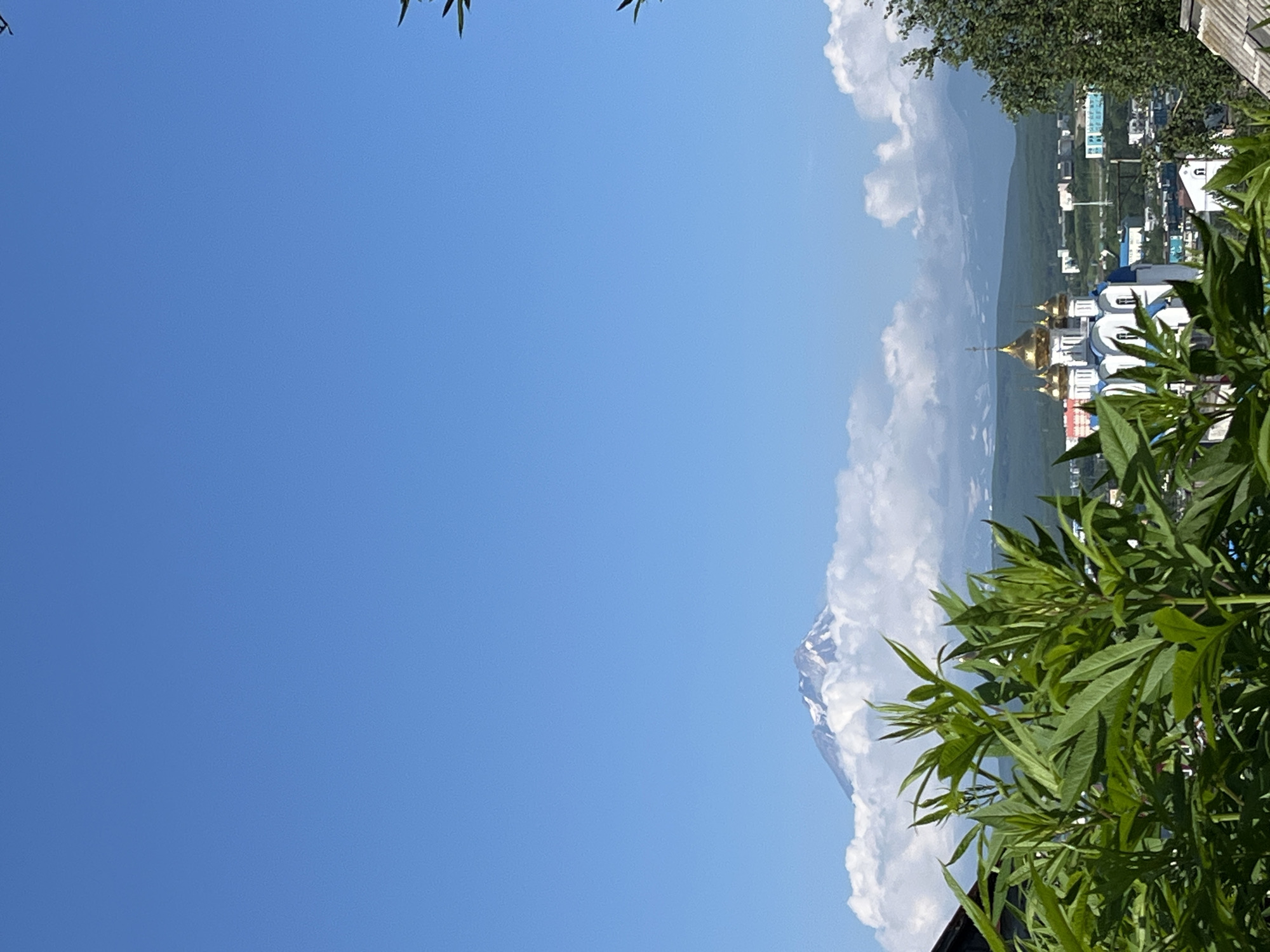 A medium shot captures a serene urban landscape in Petropavlovsk-Kamchatsky, Russia, on a clear, sunny day. In the foreground, lush green foliage partially obscures the view. Behind the greenery, a white and blue church with golden domes stands prominently, its architectural details suggesting a place of worship. The middle ground reveals a cityscape with various buildings, some appearing residential and others more commercial or institutional, with a few scattered trees. In the distance, a majestic snow-capped volcano rises above a bed of fluffy white clouds, its peak touching the clear blue sky. The overall scene conveys a sense of peace and natural beauty, blending urban development with the grandeur of volcanic landscapes.
