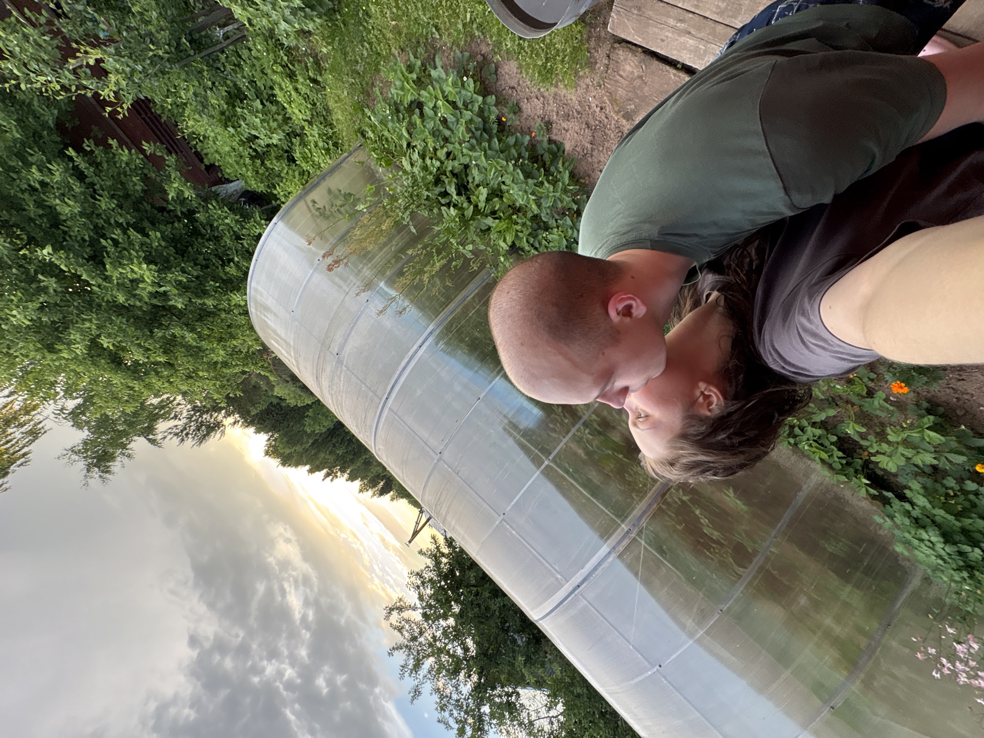 In Дресвяново, Russia, a couple shares a tender moment in front of a greenhouse during sunset. The man, bald with a shaved head, leans in to kiss the woman. She has long, dark hair and is wearing a dark t-shirt. The man is wearing a green t-shirt and blue jeans. The greenhouse, constructed with a transparent material, is a prominent feature in the background, reflecting the sky and surrounding greenery. The sky is filled with dramatic clouds illuminated by the setting sun. The scene is set in a natural environment, with lush green trees and plants surrounding the greenhouse. A wooden platform is visible in the foreground, suggesting an outdoor seating area. The overall atmosphere is romantic and peaceful.