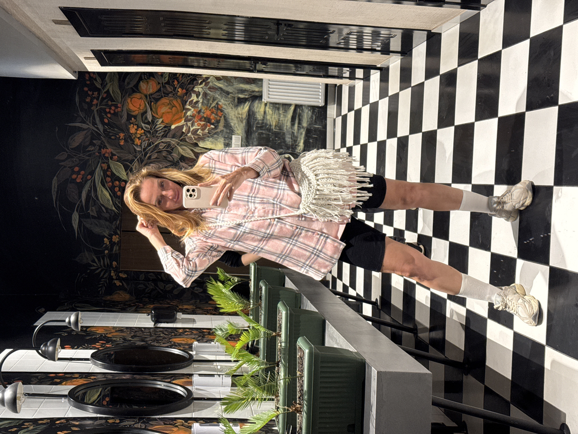 A woman stands at a slight angle in a room with black and white checkered flooring, holding a smartphone, presumably taking a selfie. She wears a pink plaid shirt, black shorts, white socks, and sneakers. A cream-colored handbag with fringes is slung across her body. Behind her, the wall is decorated with a floral and fruit mural. A row of small potted plants is positioned on a light gray countertop along with round mirrors. Above the mural, there is a narrow, horizontal vent. The location is identified as Tashkent, Uzbekistan.