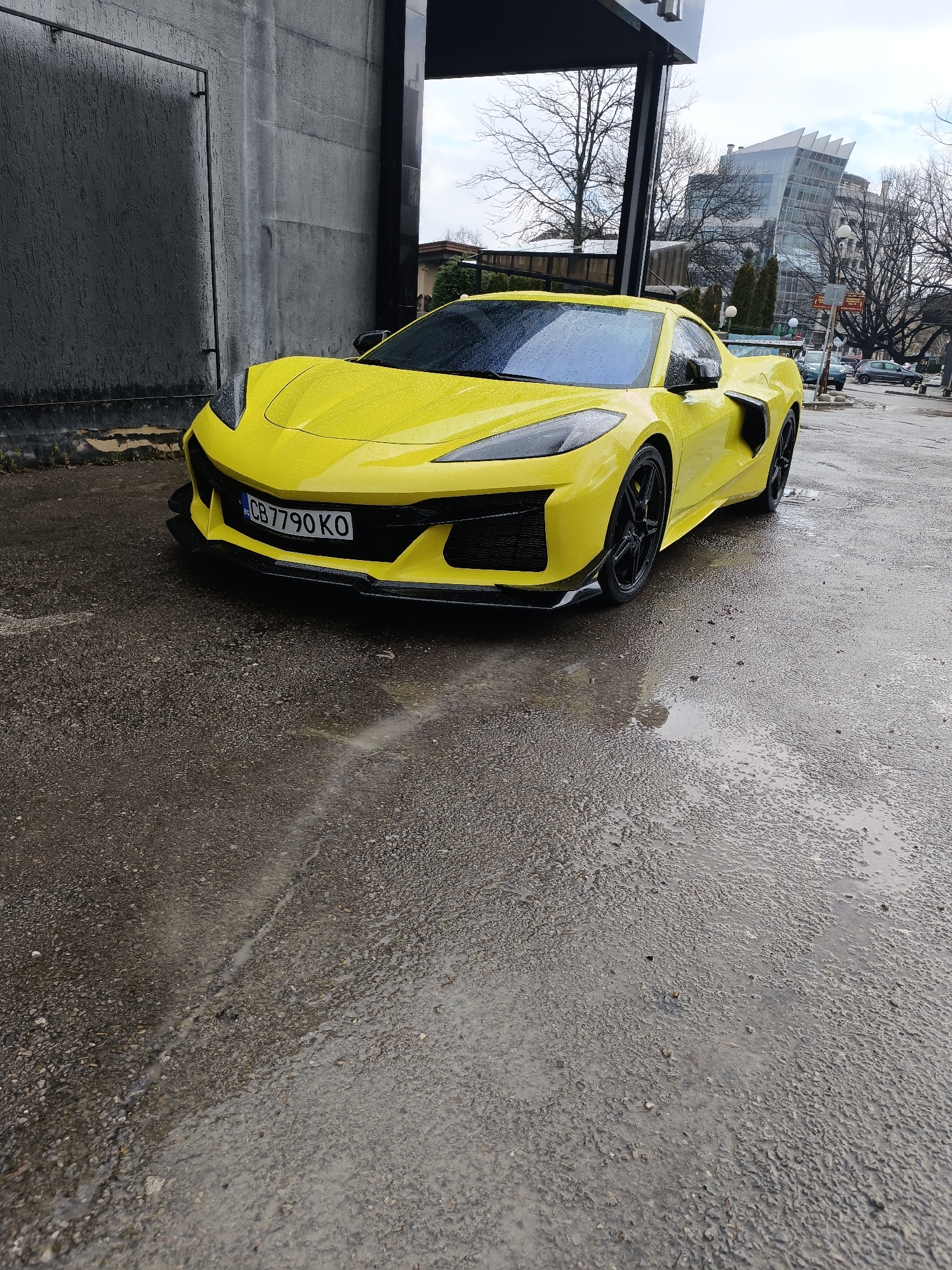 A yellow Chevrolet Corvette C8 Z06 is parked on wet pavement near a building with a black facade. The car has a Bulgarian license plate reading "CB7790KO." In the background, there is a glass building with an unusual architectural design. The environment appears to be urban, with leafless trees and other vehicles visible further down the street. The location is likely in Sofia, Bulgaria, based on the license plate.