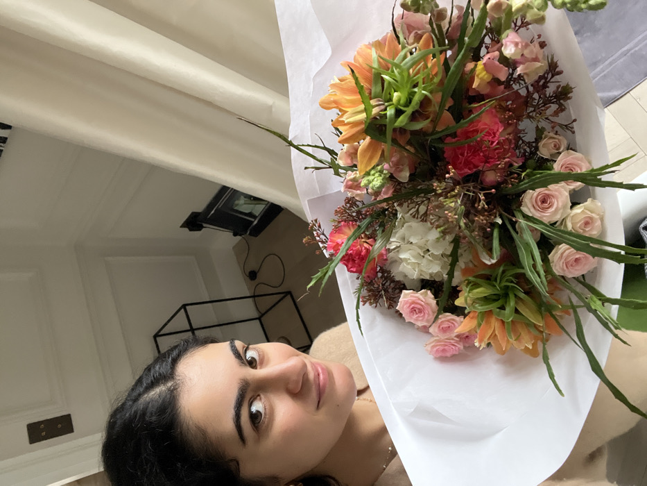 A young woman with dark hair and brown eyes is posing with a large bouquet of flowers. The bouquet has orange, pink, and white flowers wrapped in white paper. The background is a bright, modern room. A black wireframe shelf and a framed object are visible. The photo is slightly tilted and has a casual, selfie-like feel.