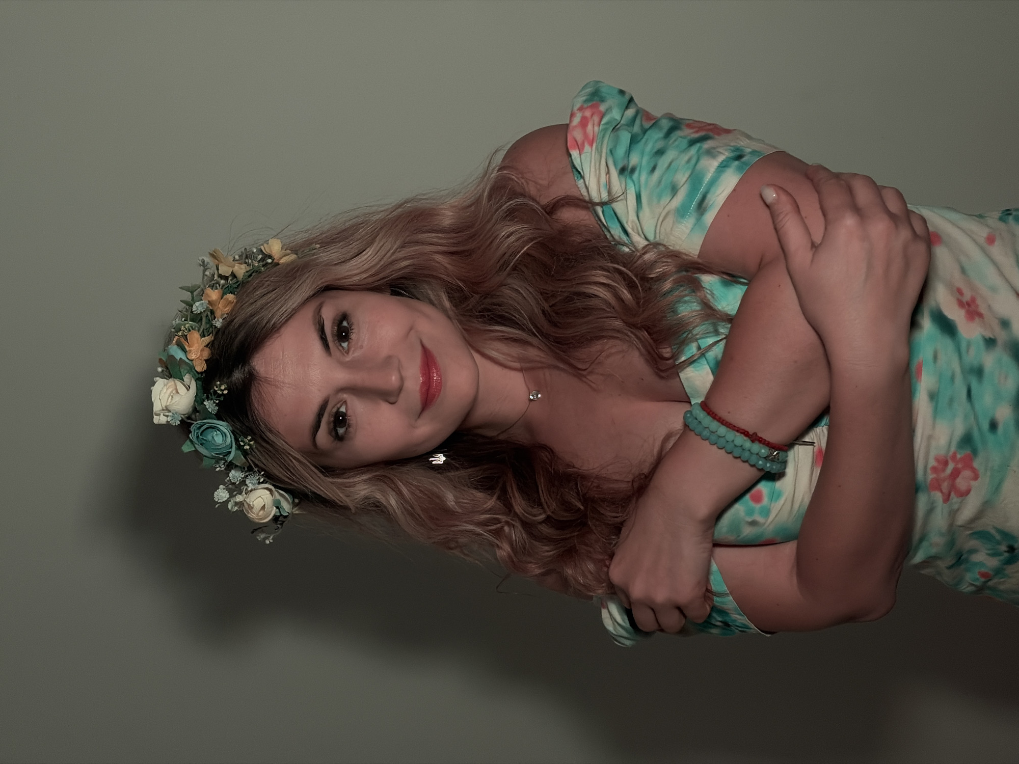 A woman with long, wavy blonde hair, adorned with a floral crown, gazes at the camera. She wears a light-colored dress with a teal and pink floral pattern, and a beaded bracelet is visible on her wrist. She has her arms crossed over her chest. The background is a plain, light gray wall. The photograph does not provide information to infer the location in Budva, Montenegro.