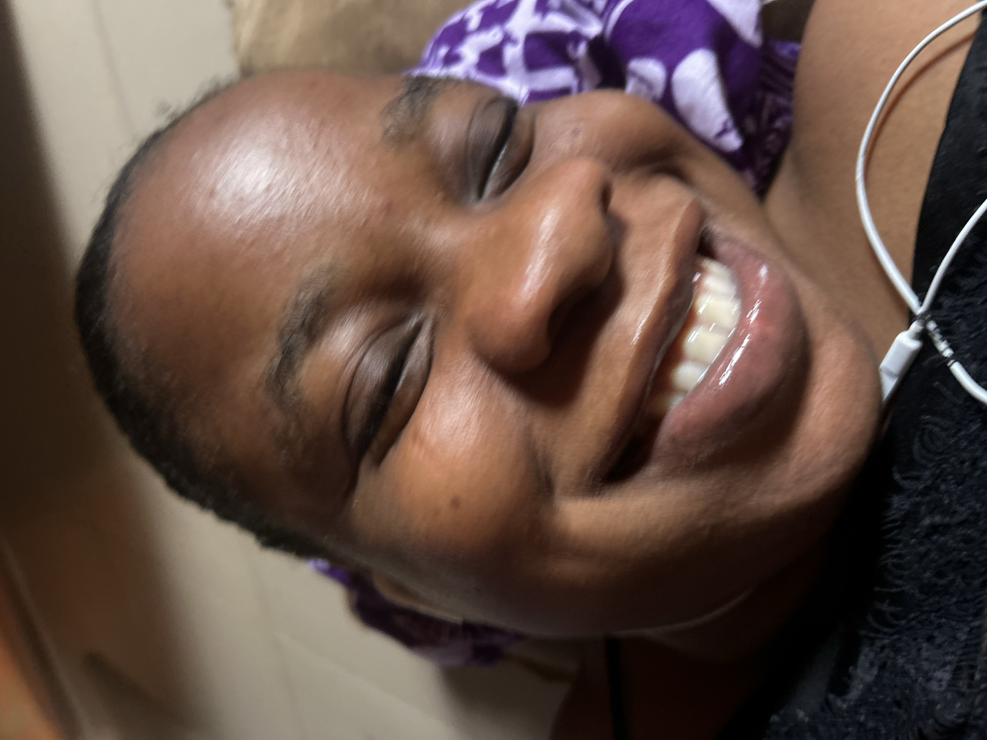 Close-up photograph of a dark-skinned woman, smiling widely with visible teeth, possibly lying down. She has short, dark hair and a shiny forehead. She wears black clothing with lace details and white wired earphones. A purple and white patterned fabric appears behind her head. The location is Unknown, Unknown.