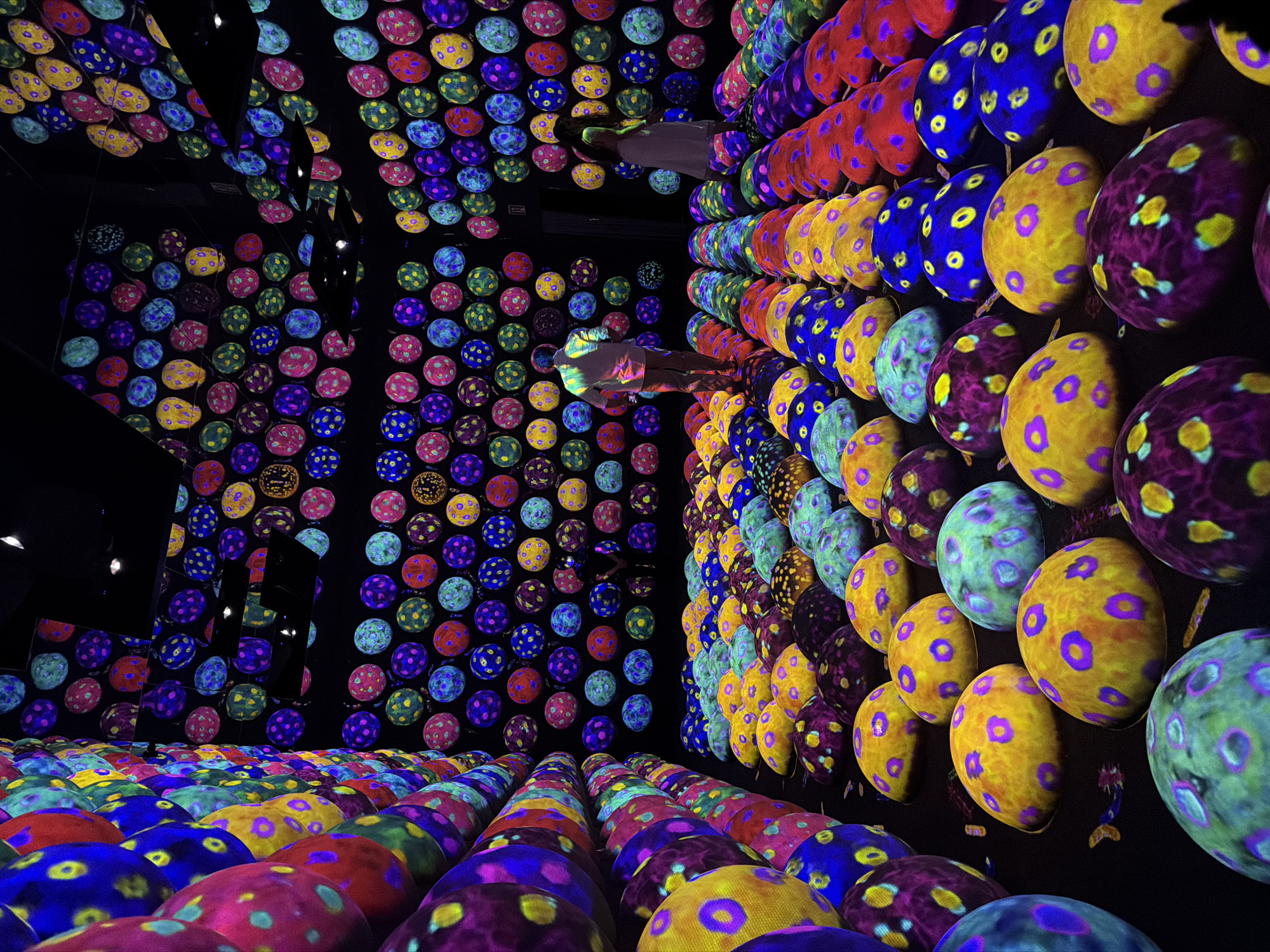 The image depicts a dimly lit, immersive art installation within the teamLab Borderless digital art museum in Koto, Japan. Rows of large, bulbous, mushroom-like sculptures, brightly illuminated with vibrant, neon-like colors and polka-dot patterns, cover the floor. These sculptures exhibit a variety of color combinations, including yellow with purple spots, blue with yellow spots, and green with blue spots. To the left, a wall is covered in more of these colorful, spherical elements, creating a vertical, textured surface. In the background, the walls and ceiling are also adorned with a grid of similar circular illuminated patterns, reflecting in mirrored surfaces to amplify the visual effect. Two individuals, a man and a woman, are visible in the middle ground, observing the installation. The man, wearing a light-colored shirt and pants, stands with his back to the viewer, facing the colorful display. The woman, in a light-colored dress, stands slightly to his right, also facing the art. The overall atmosphere is one of surreal, kaleidoscopic wonder, characteristic of digital art museums.