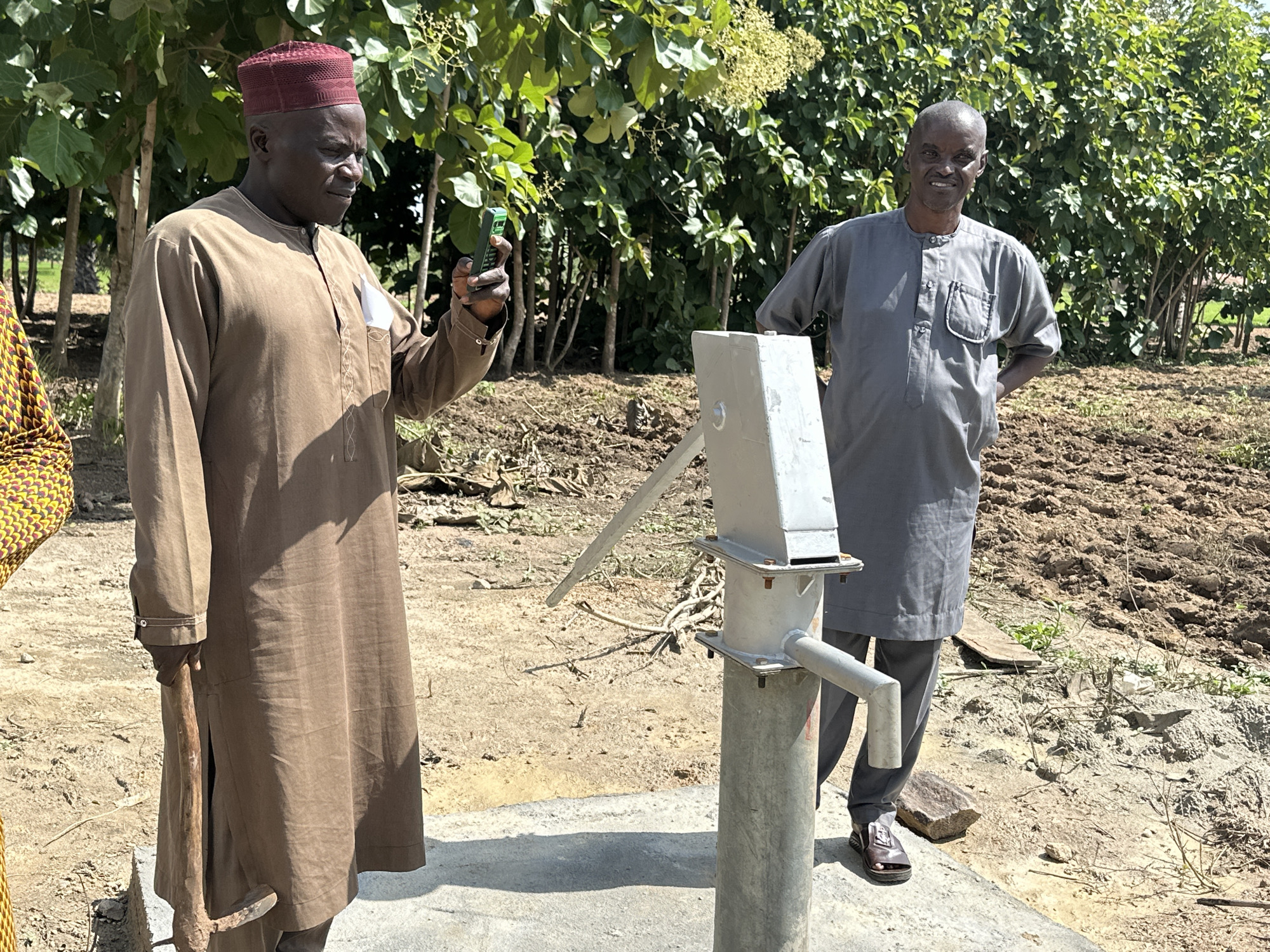 In Afogo, Nigeria, two men stand near a hand pump in a rural setting. The man on the left, wearing a brown tunic and a maroon cap, holds a green mobile phone. The man on the right, in a grey tunic, stands with his hands behind his back. The hand pump is a metallic structure with a long handle. The ground is dry earth, with scattered vegetation and a backdrop of lush green trees. The scene appears to be under bright daylight, suggesting an outdoor location.