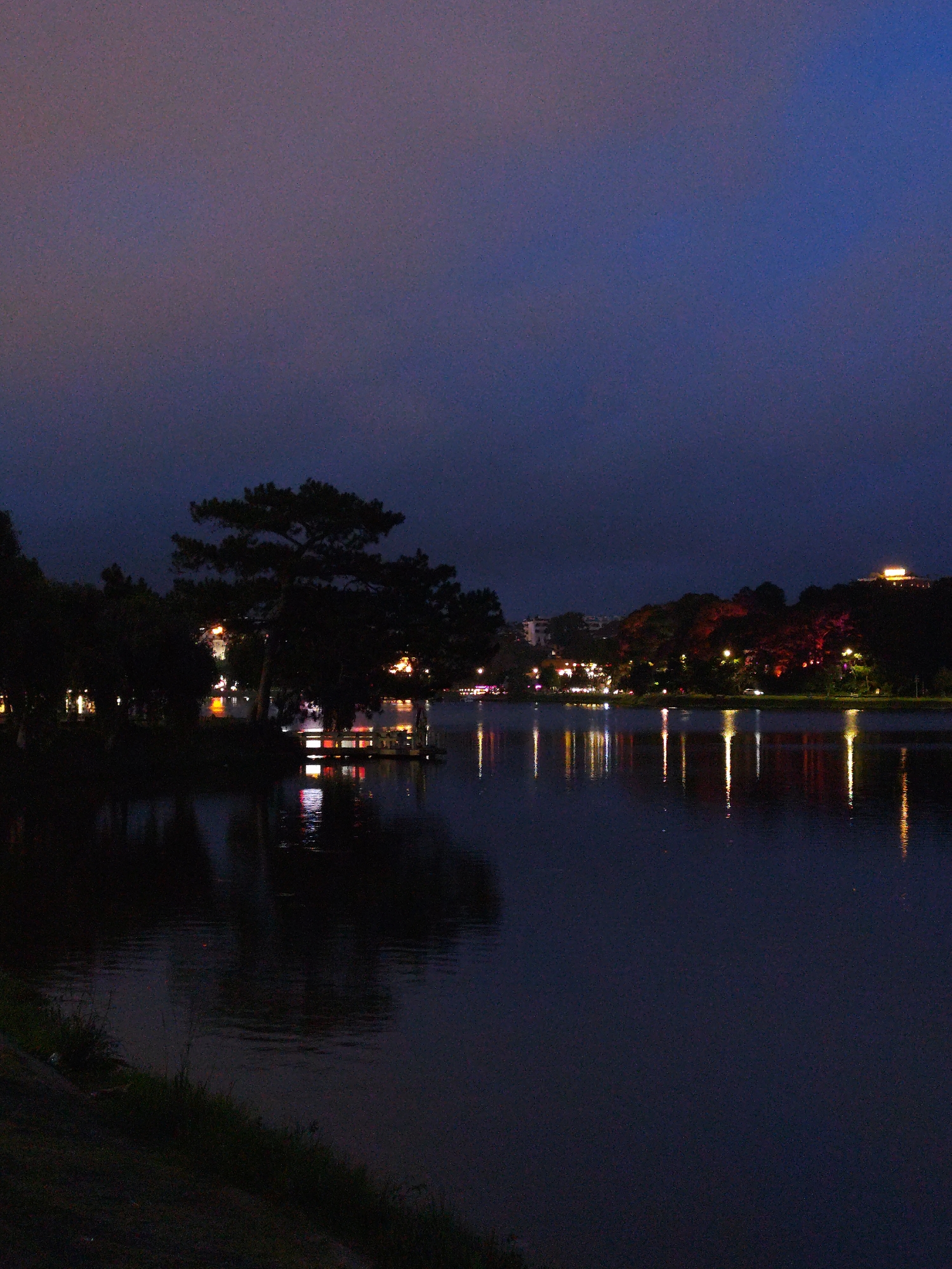 At night, a body of water, possibly a lake, is the focal point, its dark surface reflecting the lights on the distant shore. These reflections form vertical lines of varying brightness and color. The sky above is a gradient of dark blue and purple, suggesting dusk or late evening. A dark silhouette of a large tree stands prominently on the left, partially obscuring a lit structure. On the right shore, a row of trees is illuminated with red and white lights, and a building with a brightly lit roof is visible on a hill in the background. The foreground shows a dark shoreline with some vegetation. This scene is likely located in Da Lat, Vietnam, known for its lakes and hilly landscapes, as suggested by the surrounding environment.