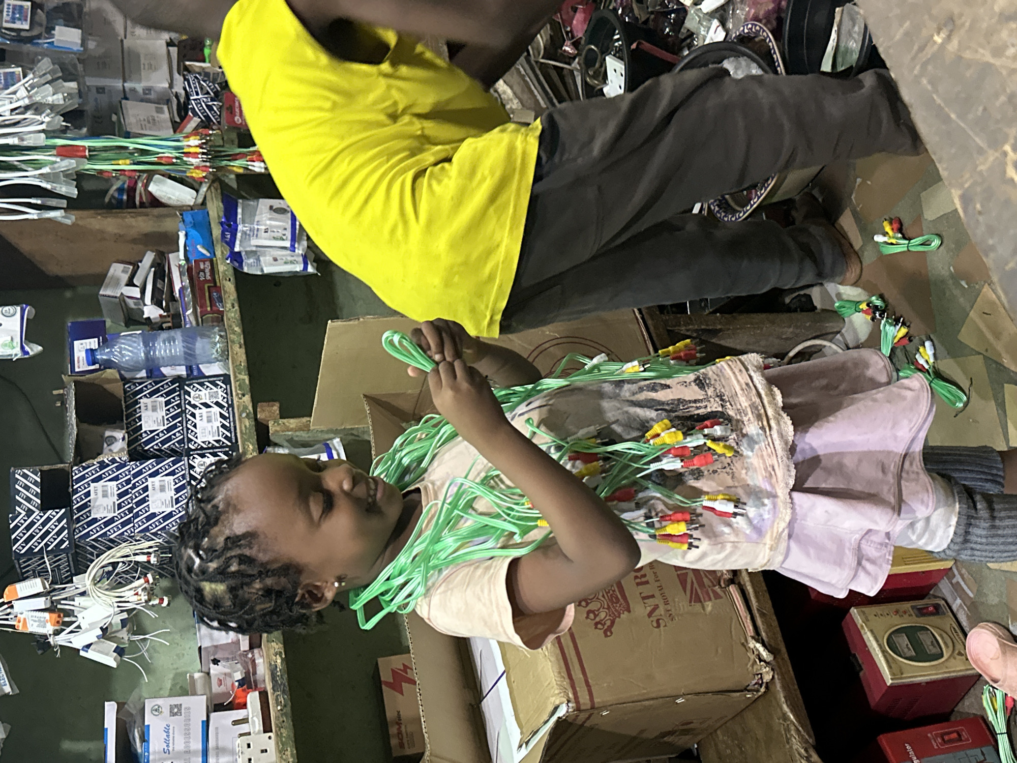 In Narayi, Nigeria, a young girl with braided hair is seated on the floor, surrounded by a chaotic array of electronic components and cables. She is holding a bundle of green audio/video cables, her face illuminated by a bright, natural light, suggesting it is daytime. The setting appears to be a workshop or storage area, with stacks of cardboard boxes, some containing electronics and others labeled with text. One box shows the text "AFFET." Another box displays "SNT ROYAL FOR V." In the background, an adult wearing a bright yellow t-shirt and dark pants is partially visible, also surrounded by the electronic clutter. The girl's expression is one of focus and engagement with the cables. The overall scene conveys a sense of industrious activity and the potential for repair or assembly of electronic devices.