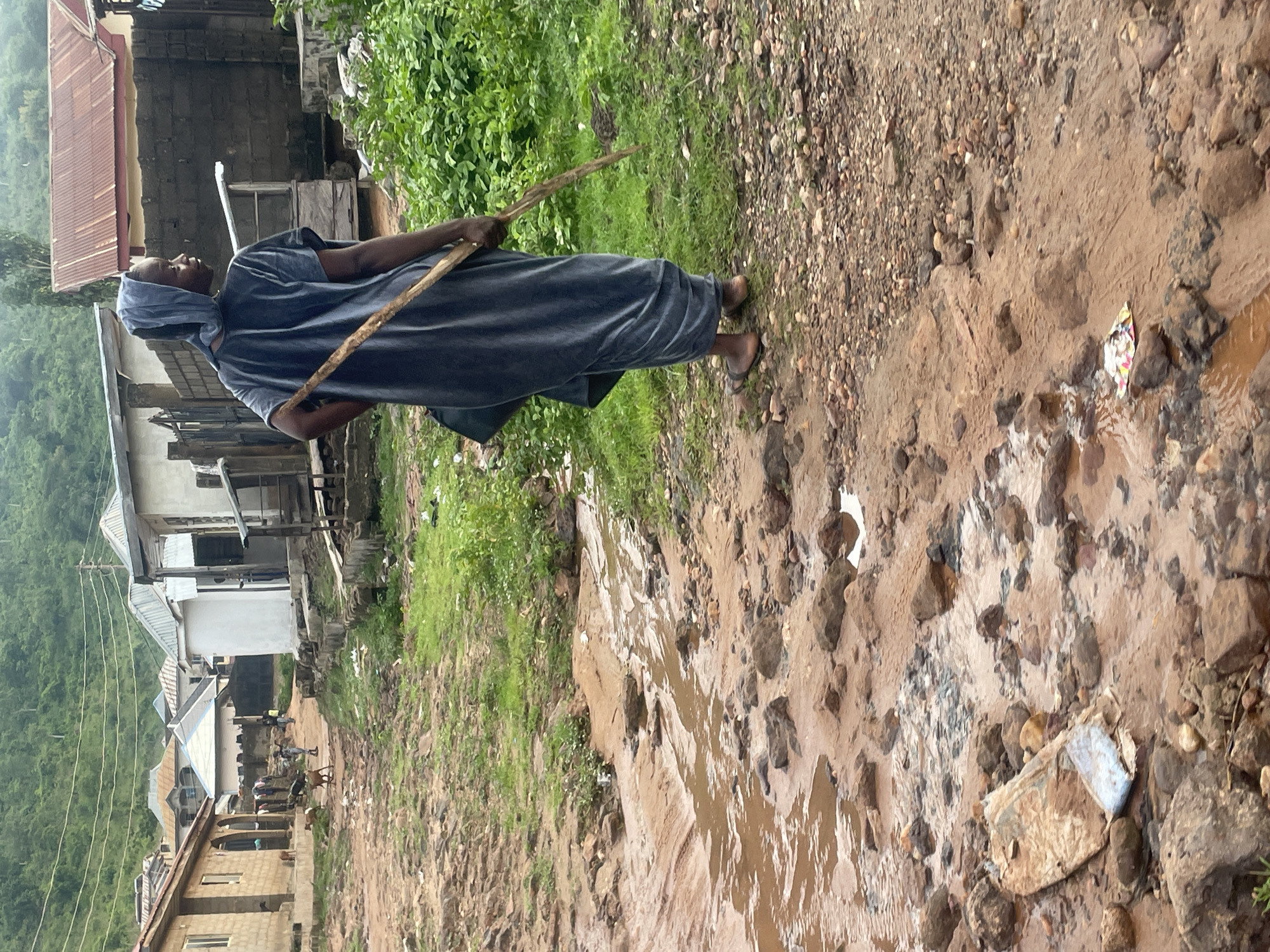 The image depicts a scene in the city of Lokoja, Nigeria. In the foreground, a dirt path is partially flooded with muddy water, with rocks and debris scattered throughout. The path leads towards a settlement of buildings with corrugated metal roofs and concrete walls.
A person, likely a woman given the attire, is seen walking away from the camera on the right side of the path. She is wearing a dark blue, loose-fitting garment that covers her head and body, and sandals. She is carrying a long, light-colored wooden stick over her right shoulder.
In the background, several residential buildings are visible, along with more greenery and a hilly landscape. A few animals, possibly goats, are present on the path near the buildings in the mid-ground. The overall environment appears to be rural or semi-urban, with evidence of rain or recent rainfall due to the muddy conditions.