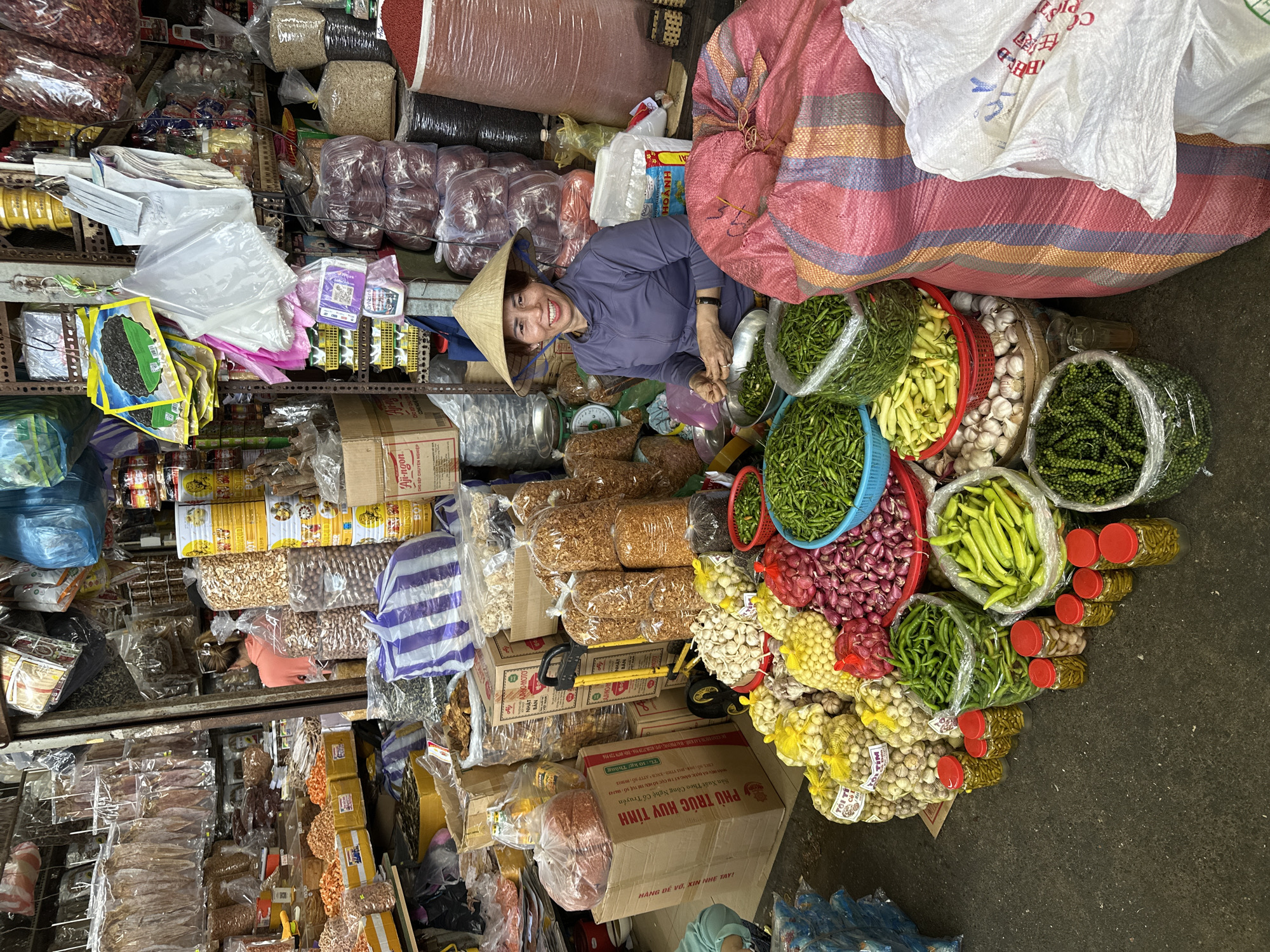 This image captures a vibrant market stall in an indoor setting, identified as located in Đà Nẵng, Vietnam. The scene is densely packed with merchandise, featuring a central female vendor engaged in preparing produce.
The primary subject is an adult woman positioned slightly left of center, wearing a purple long-sleeved top and a traditional conical straw hat (nón lá). She is smiling directly at the viewer. Her hands are occupied with a small metal bowl containing green chilies, suggesting she is sorting or preparing them for sale.
In the immediate foreground, a wide array of fresh produce is displayed. Piles of red shallots or small onions, large quantities of garlic bulbs (some packaged in yellow mesh bags labeled "TỎI TIM"), and various types of green chilies are arranged in colorful plastic baskets and clear plastic bags. Several small clear glass jars with red lids, containing what appears to be pickled chilies or green peppercorns, line the concrete floor. A large white bag and a red and multi-striped woven bag, both filled with goods, are positioned on the right side of the frame. A plastic bag brimming with green peppercorns is also visible.
Behind the vendor, a cluttered market stall extends upwards and into the background. Shelves are laden with numerous packaged goods. These include stacks of clear plastic bags filled with various nuts (cashews, peanuts) and dried shrimp. On the far left, clear plastic packages containing dried squid and other dried seafood hang from hooks. Yellow canned goods are visible behind the nut bags. Further back on the right, large sacks of grains or beans, including red beans, are piled high. Various cardboard boxes, some labeled "AJI-NGON" (Ajinomoto) and "PHÚ TRÚC HUỲNH," are stacked throughout the stall. A green weighing scale with a white face is visible behind the vendor. Overhead, numerous plastic bags of different colors (blue, white, pink) are hanging, along with packages of dark, dried food items, possibly seaweed or fungi. A yellow hand truck is partially visible in the mid-background. The overall environment is characteristic of a bustling, densely stocked local market.