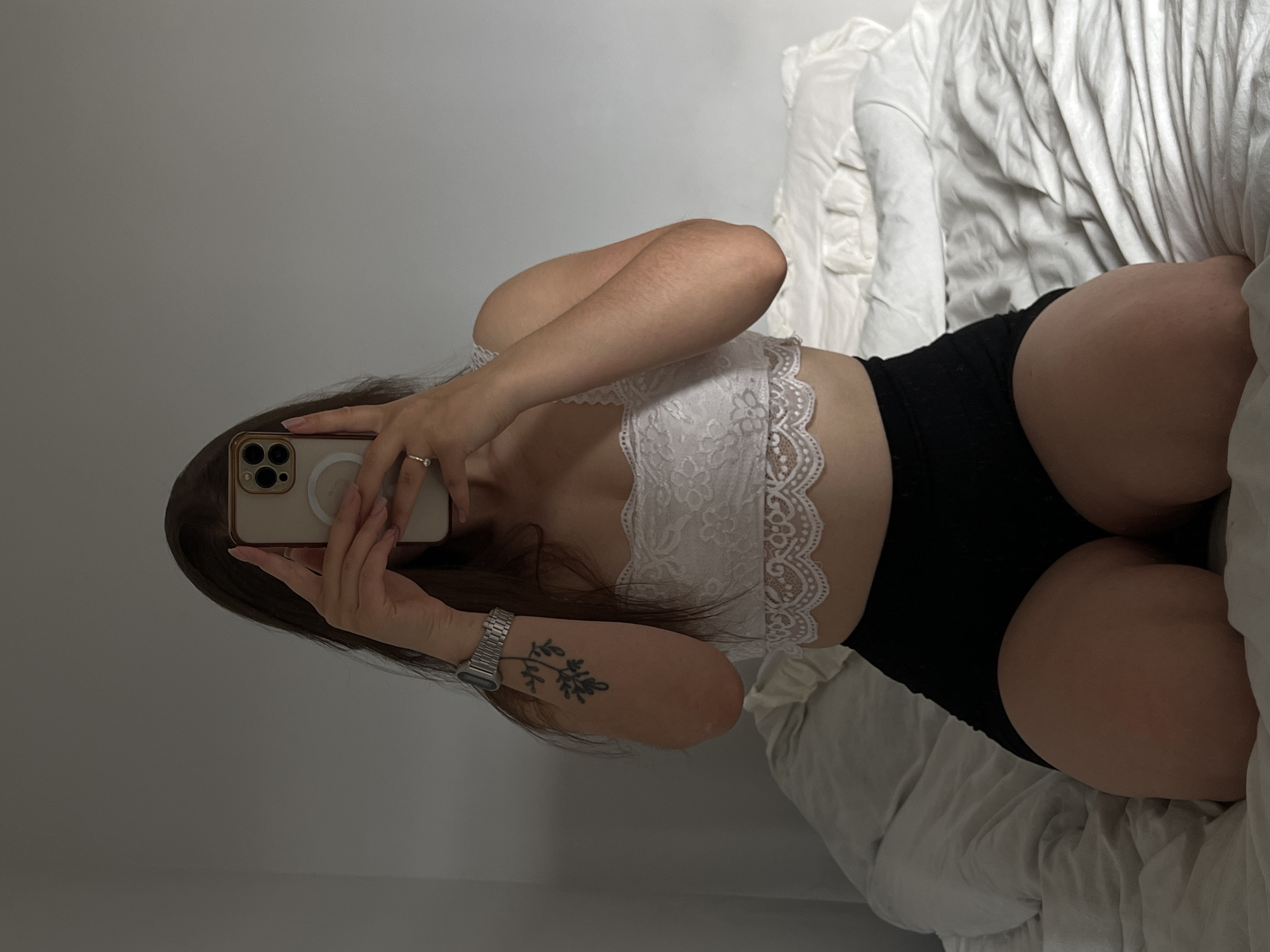 A woman is sitting on a bed, holding a smartphone to take a picture of herself in a mirror. She is wearing a white lace bralette and black shorts. The bed is covered in white sheets and pillows. The background is a plain white wall. There is no indication of the city of Ternopil, Ukraine in the image.