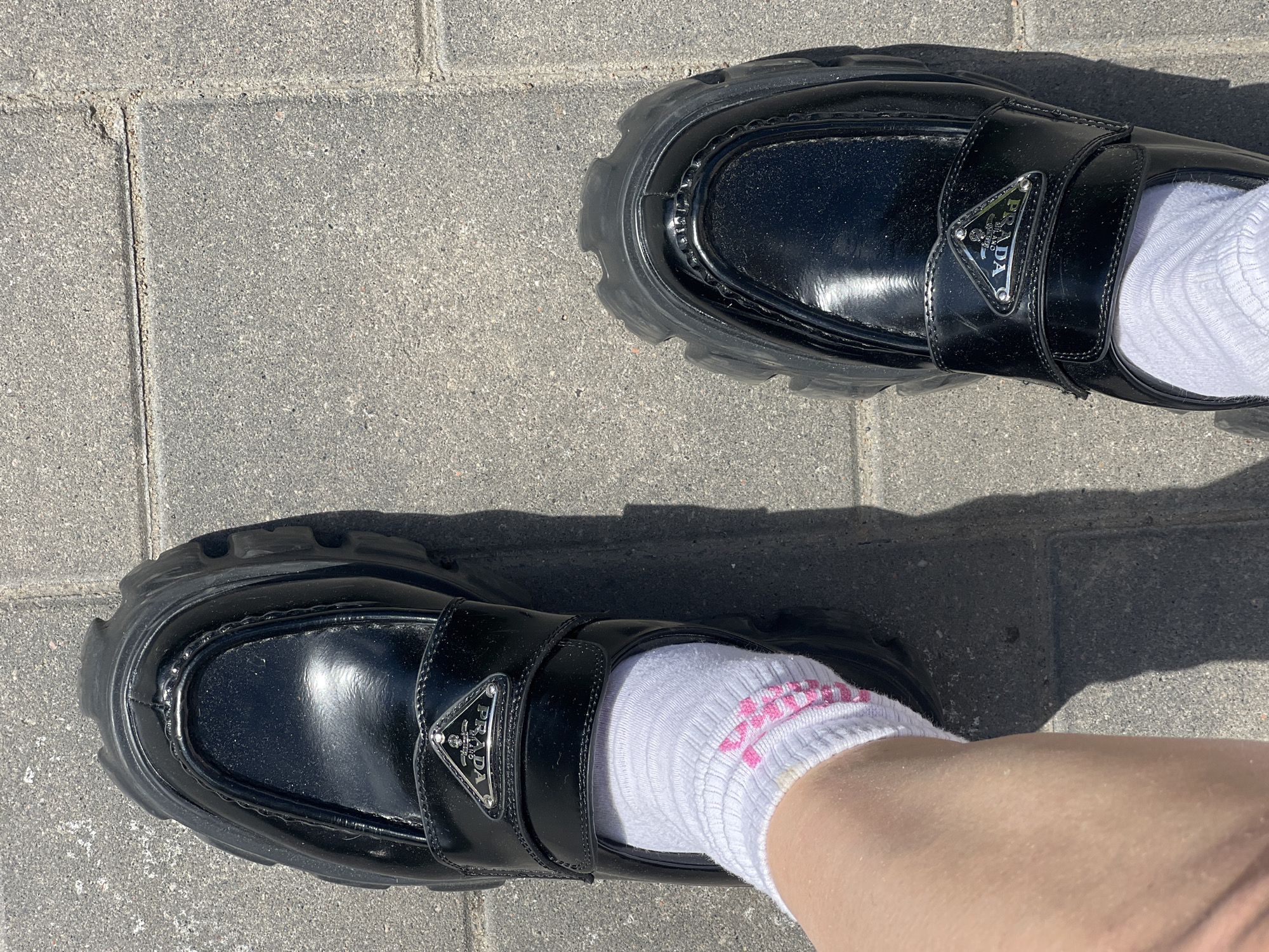 A pair of black patent leather Prada loafers with thick rubber soles and a silver Prada logo are on a person's feet, who is wearing white socks with pink stripes. The person is standing on a paved walkway made of light gray rectangular stones. The scene is outdoors, with a shadow cast by the person and their shoes on the walkway, indicating a sunny day. The location is a city in Russia, but the specific city is unknown.