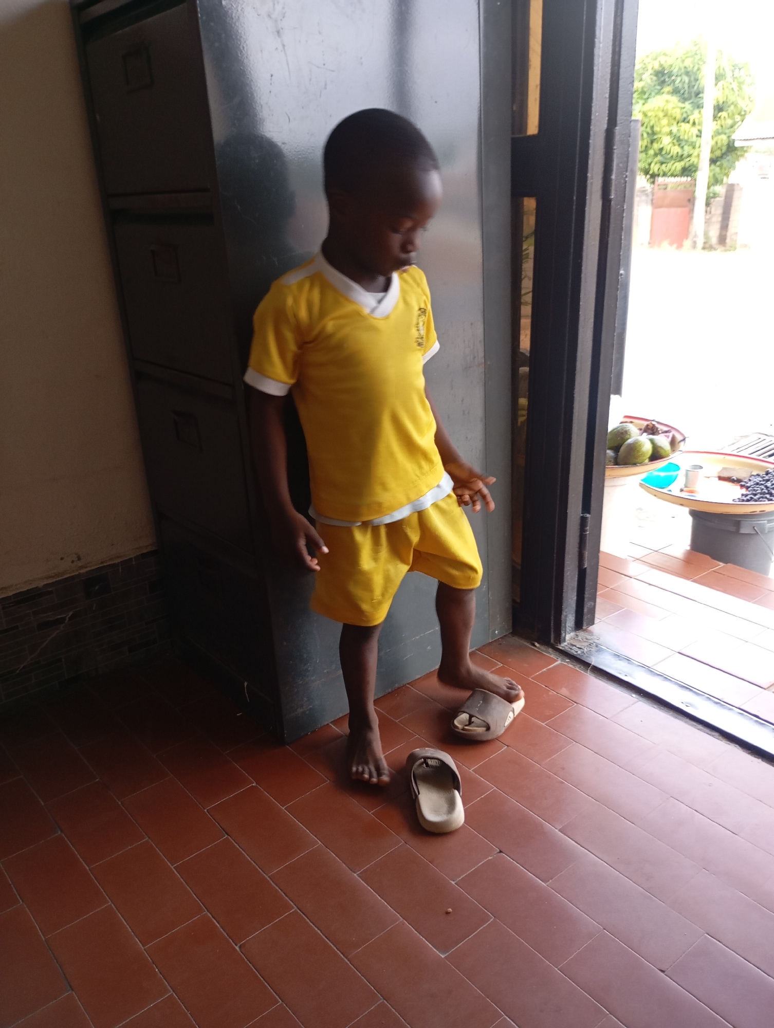 A young boy stands in a doorway, his body angled slightly away from the camera. He is wearing a yellow t-shirt with white trim and yellow shorts. His feet are bare, and he is standing next to a pair of brown and white slide sandals. The floor is tiled with reddish-brown rectangular tiles. To the left of the boy is a tall, dark gray metal cabinet. The doorway opens to an outdoor scene with trees and a building visible in the background. In the foreground of the outdoor area, on a lower level, are two shallow bowls filled with fruits, one with green avocados and the other with dark berries, and a small metal container. The scene appears to be in Jos, Nigeria, given the presence of avocados and the general environment.