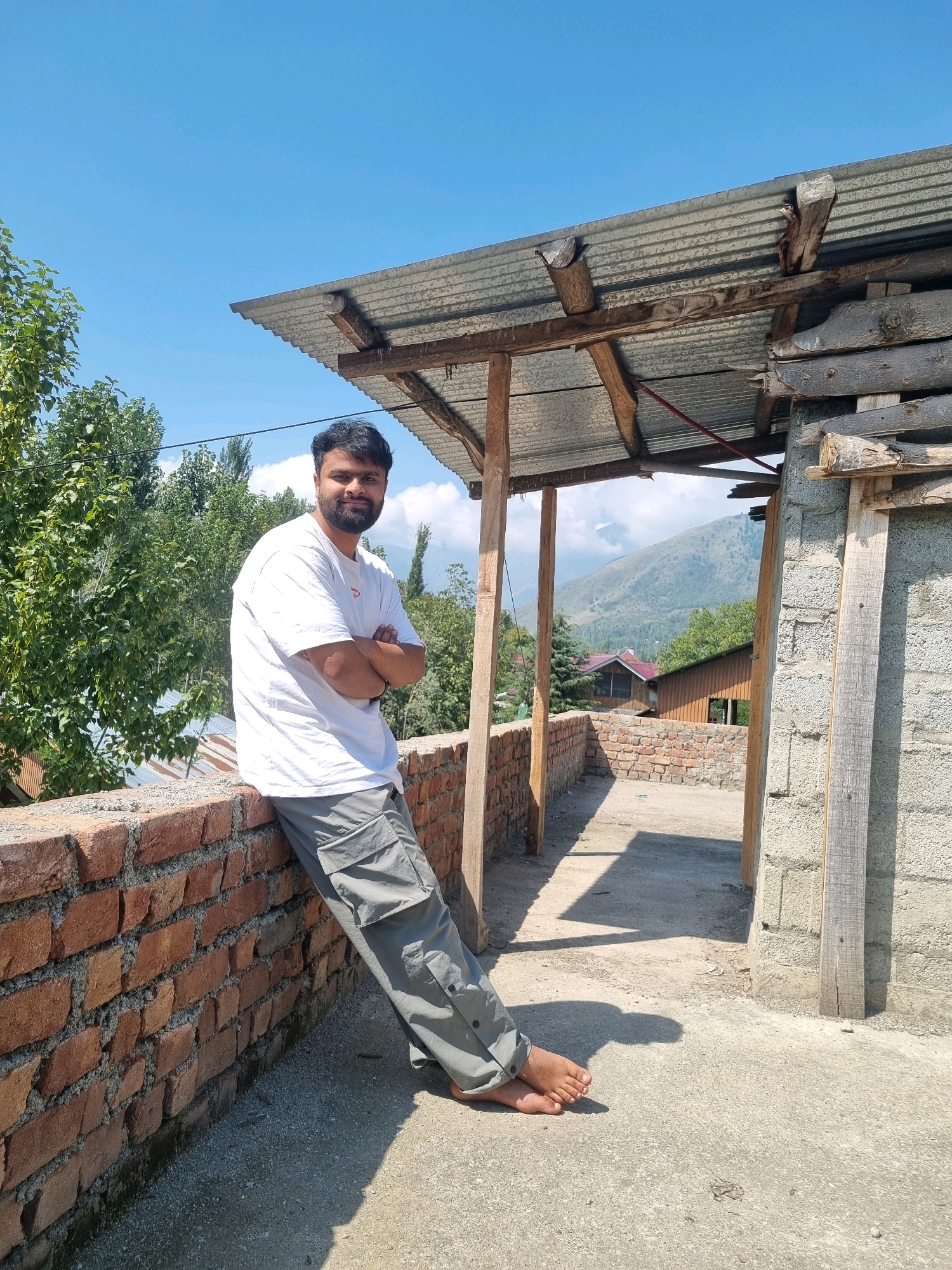 In Manigam, India, a man stands outdoors, leaning against a brick wall. He is wearing a white t-shirt and grey cargo pants, with his arms crossed. His bare feet are visible as he leans, his body angled slightly towards the camera. The background reveals a rural landscape with trees and mountains under a clear blue sky. To the right of the man, a rudimentary structure with a corrugated metal roof supported by wooden beams is visible, indicating an outdoor or under-construction area. The ground is a mix of concrete and gravel.