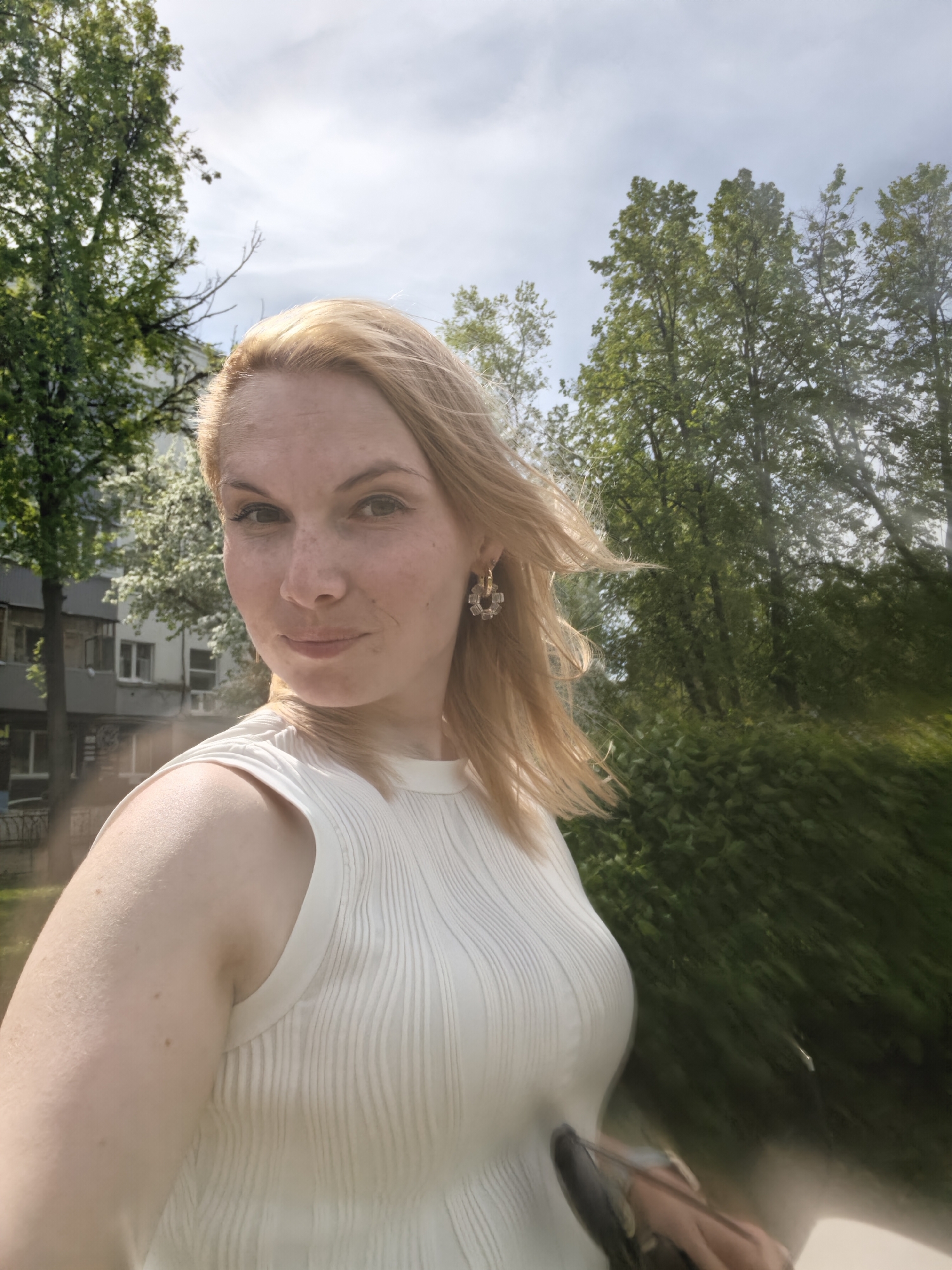 A fair-skinned woman with blonde hair is pictured outdoors. She wears a sleeveless white ribbed top and is adorned with earrings. Her expression is neutral with a slight smile.
Behind her, foliage suggests a park or garden setting. A multi-story building is partially visible to the left, lending to the urban context. Motion blur on the right implies movement or a dynamic shot. The photograph, presumably a selfie, appears to be taken in Sterlitamak, Russia, based on the prompt’s request. The lighting indicates a bright, sunny day.