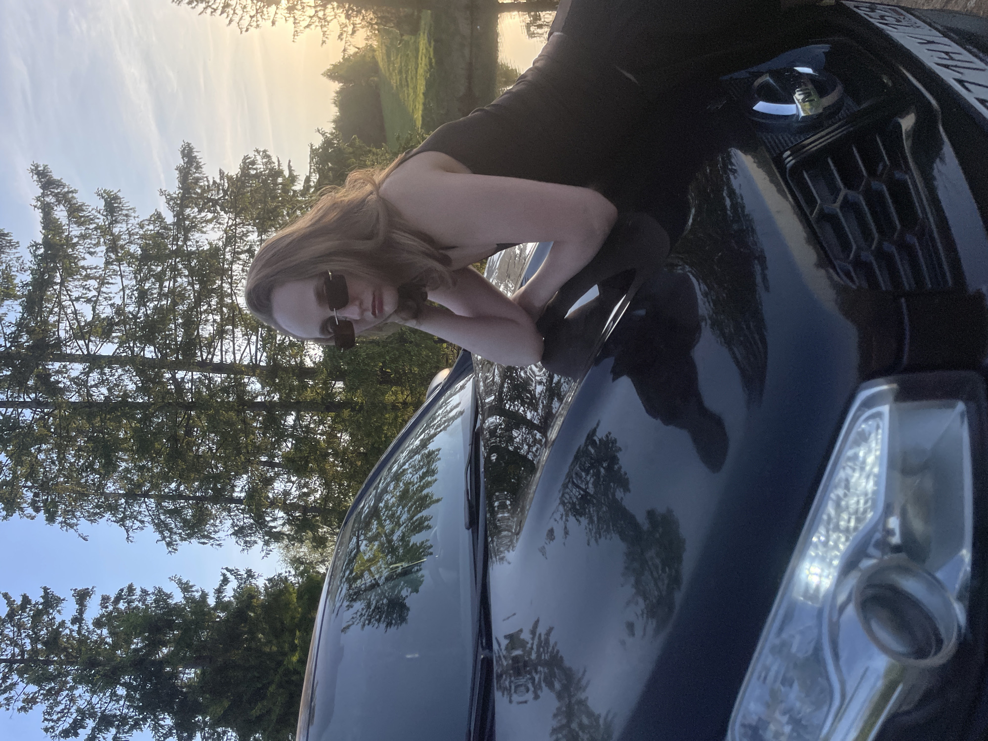 A woman with long brown hair and sunglasses, wearing a black dress, is lying on the hood of a dark-colored car. She is resting her forearms on the hood. The car is parked outdoors near trees and possibly a body of water, as suggested by the reflected light. The time of day appears to be dusk or dawn, based on the lighting. There is no definitive information to pinpoint the exact location as Подшивалово, Russia; therefore, the presence of the city cannot be confirmed.
