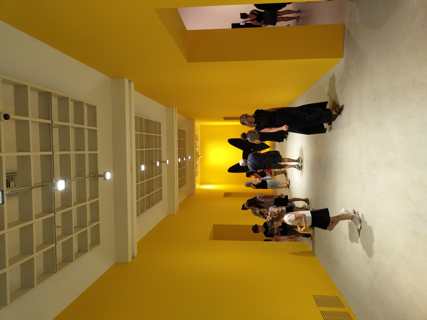 The image depicts an interior view of the Tel Aviv Museum of Art's Herta and Paul Amir Building. The scene shows a brightly lit, contemporary exhibition space with vibrant yellow walls. In the foreground, a young girl with long, curly brown hair is walking to the left, wearing a white t-shirt and a black skirt. She is casting a shadow on the light grey floor. Several other people are visible in the background, appearing to be visitors in the museum. They are dispersed along the corridor, some walking and some standing and observing. A large, black, moth-like silhouette is mounted on the far yellow wall. The architecture features a high ceiling with a grid-like structure and recessed lighting. The overall impression is that of a modern art museum in Tel Aviv, Israel.